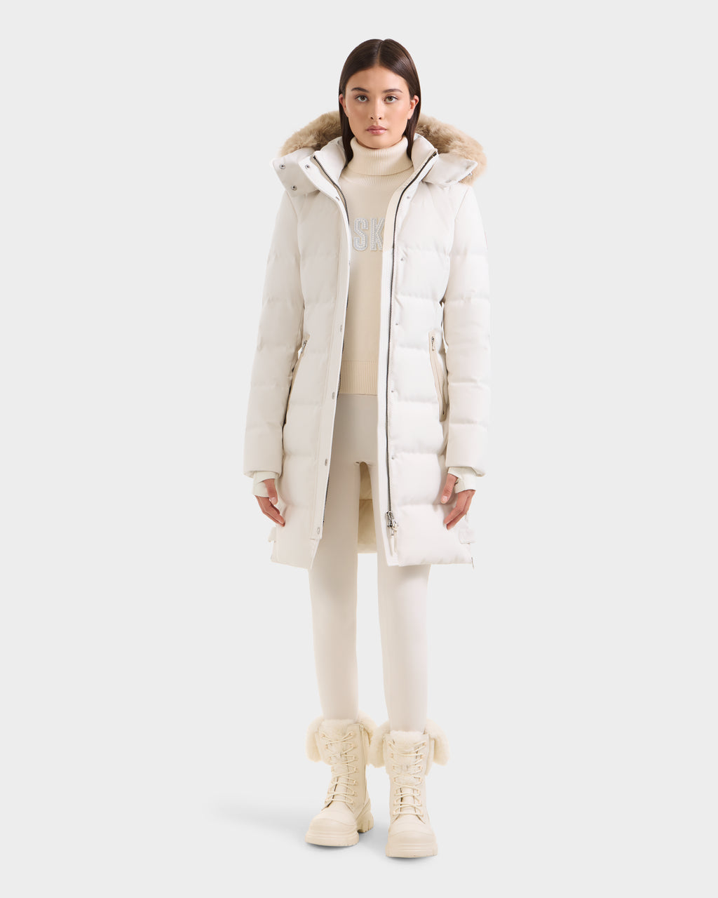 A woman faces forward in the Rudsak KEHLANI H - 8125643 Vanilla women’s down parka, a long white puffer with fur-lined hood, styled with a cream sweater, matching leggings, and fur-trimmed cream boots against a plain background.