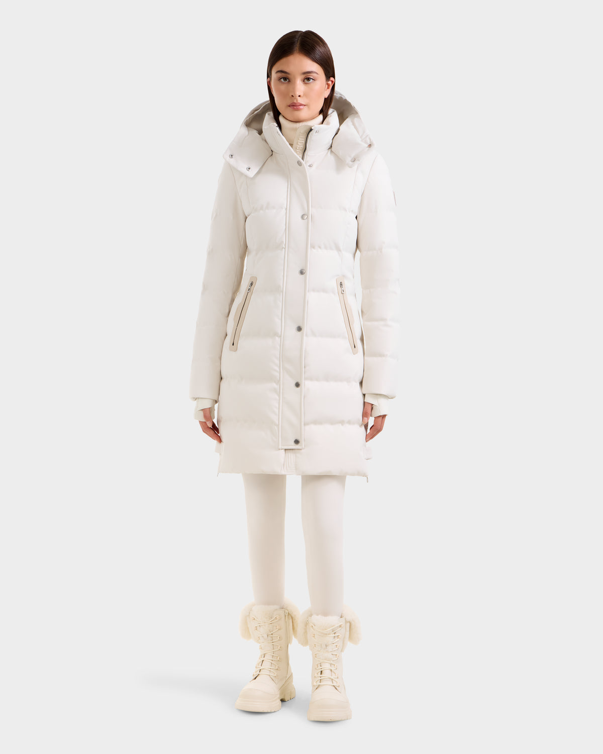 A woman stands facing forward in a long white Rudsak KEHLANI H - 8125643 Vanilla women’s down parka with RDS-certified down, white leggings, and cream fur-trimmed lace-up boots, against a plain light background.