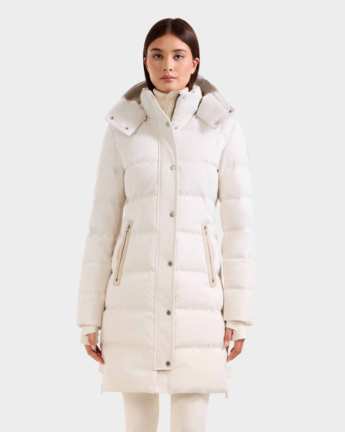 A woman stands facing forward with arms relaxed, wearing the Rudsak KEHLANI H - 8125643 Vanilla long white down parka, featuring a high collar and zippered pockets, against a plain light background.