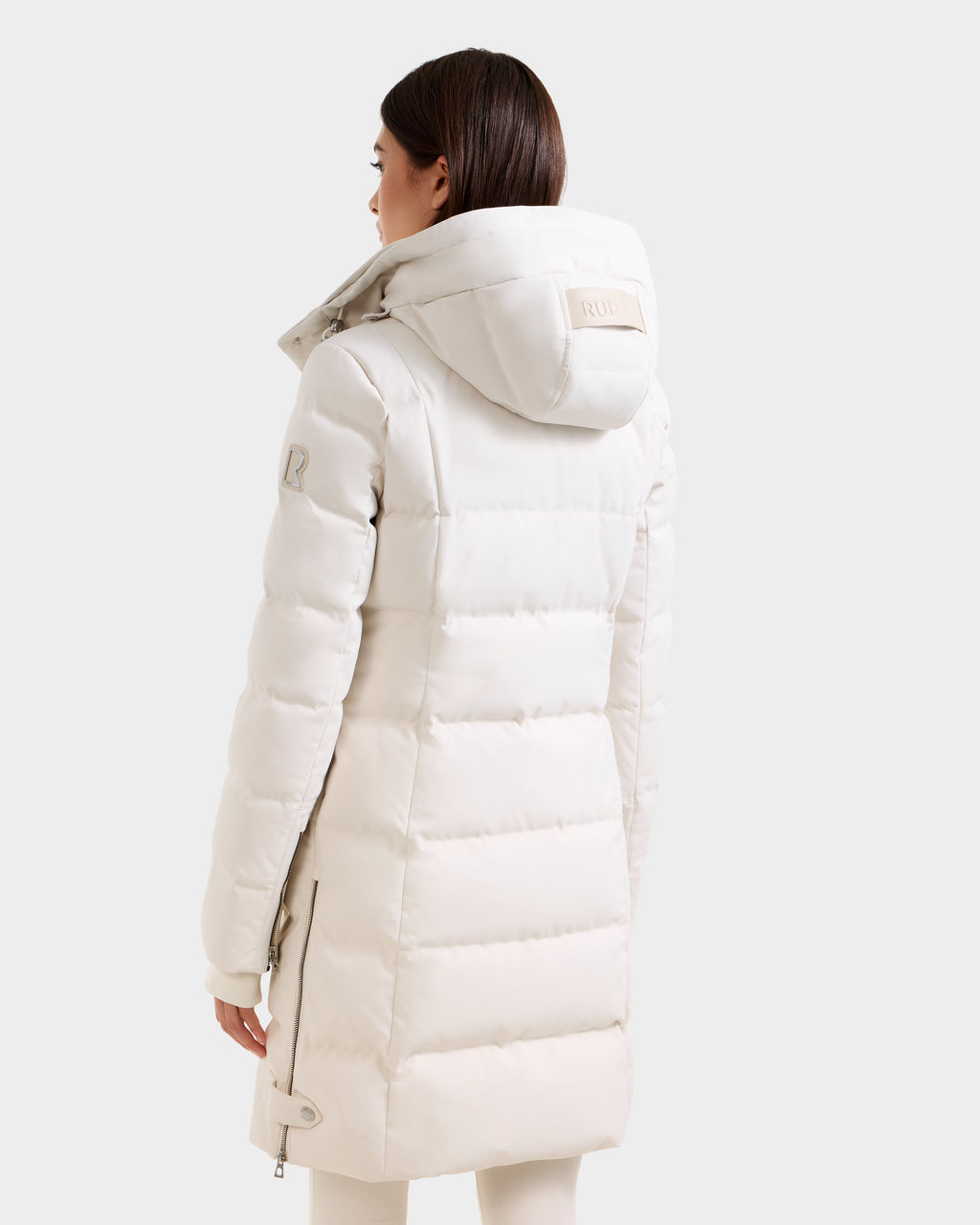 A woman with straight brown hair wears the Rudsak KEHLANI H - 8125643 Vanilla women’s down parka with hood, facing away to show the back and side of this cozy RDS-certified winter coat against a plain light background.