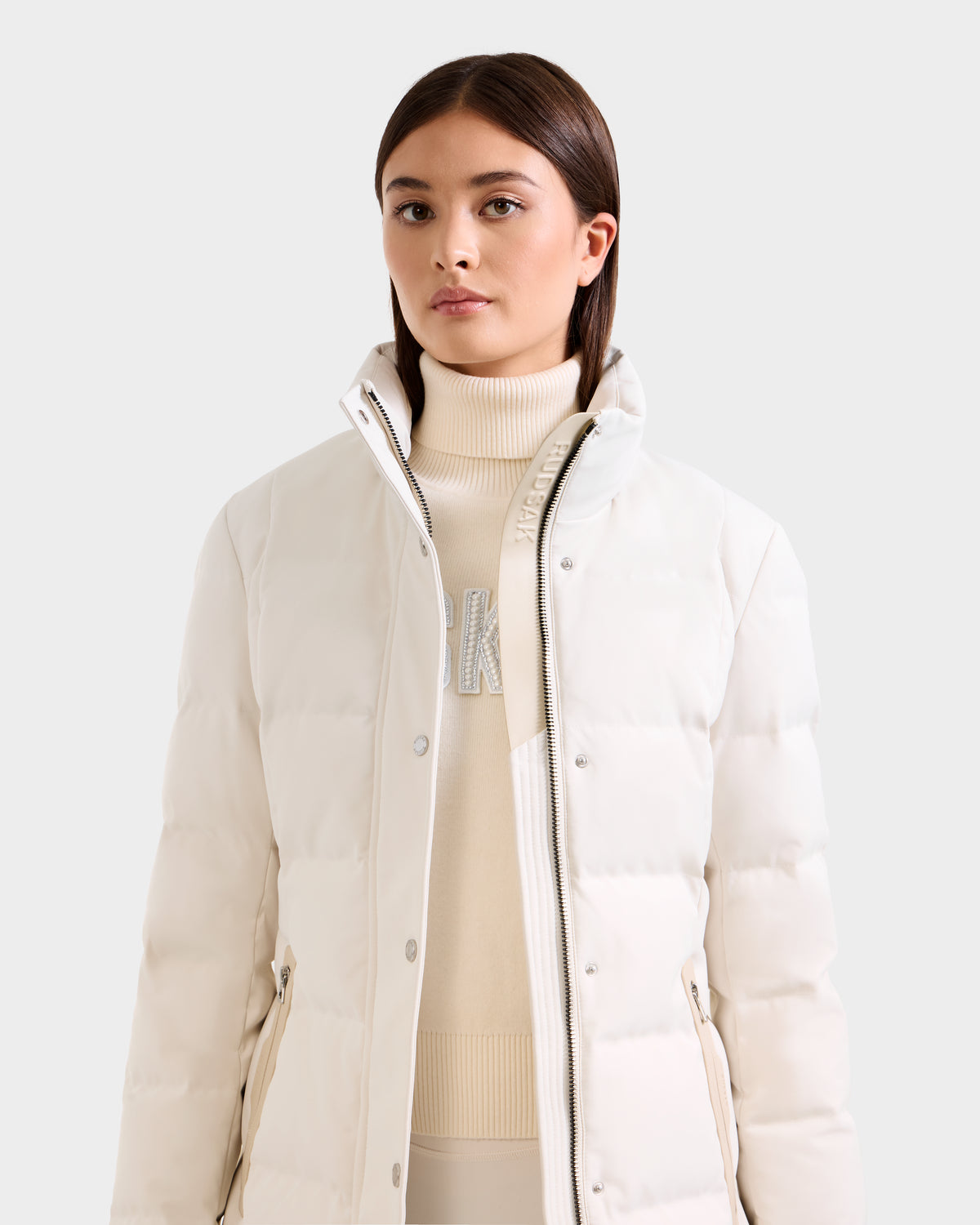 A woman with straight brown hair models the Rudsak KEHLANI H - 8125643 Vanilla women’s down parka over a white turtleneck sweater with lettering, standing against a plain light background.