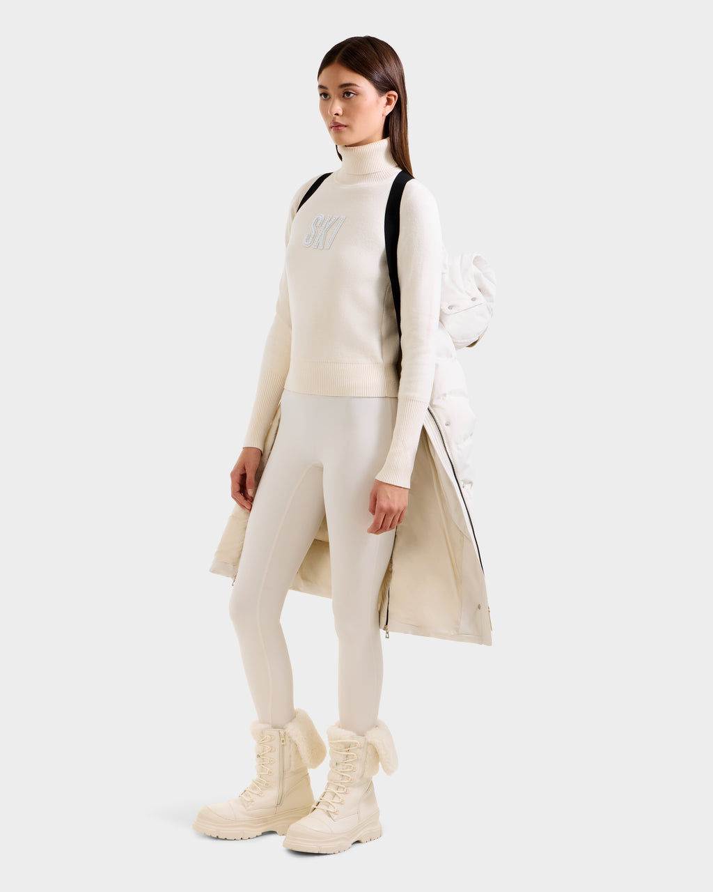A woman faces slightly to the side, wearing an all-white winter look with a long-sleeve turtleneck, fitted pants, fluffy boots, and a Rudsak KEHLANI H - 8125643 Vanilla down coat draped over her back like a cape.