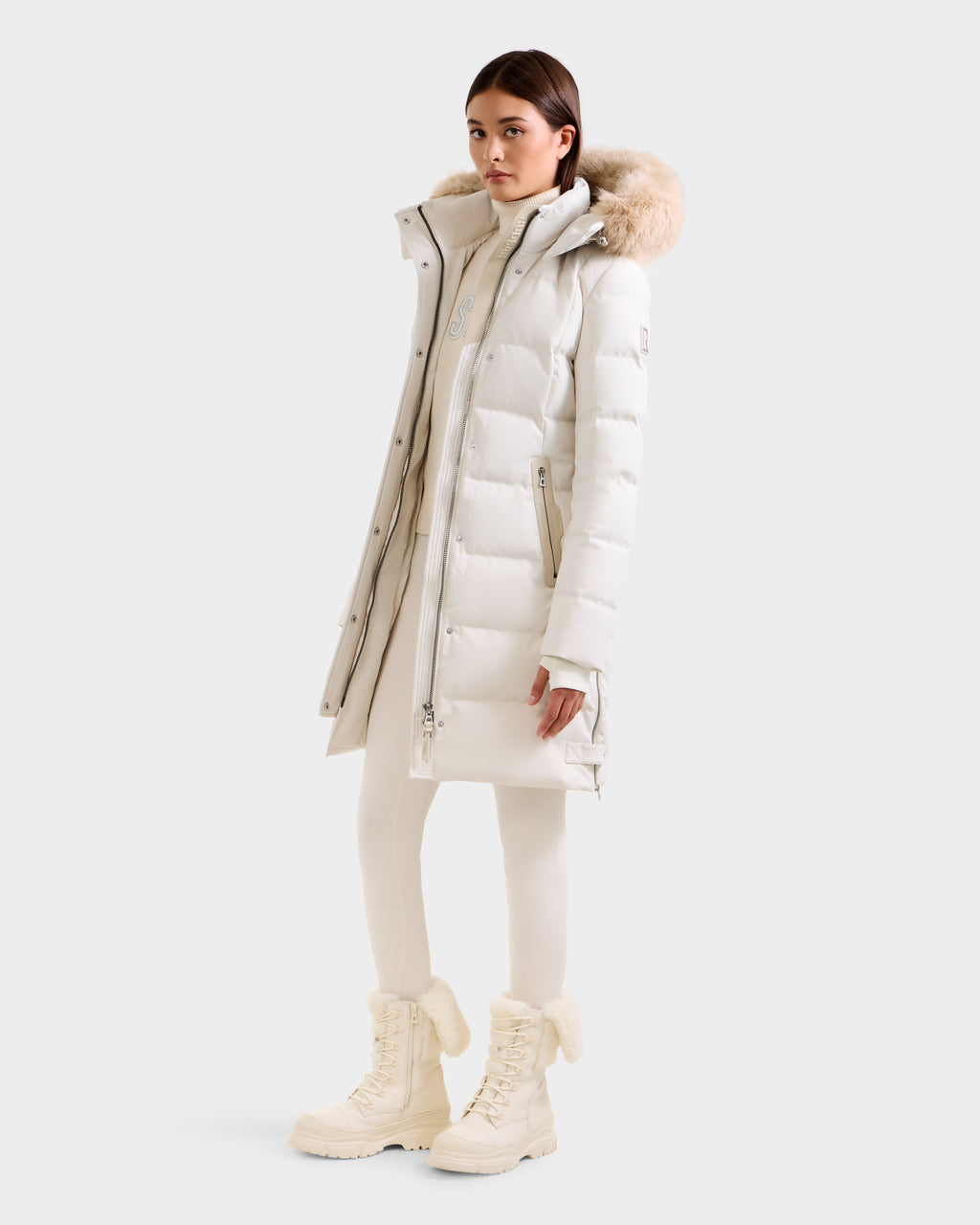 A woman wears the Rudsak KEHLANI H - 8125643 Vanilla long white down parka with a fur-lined hood, paired with white pants and winter boots, standing slightly to the side against a plain light background.