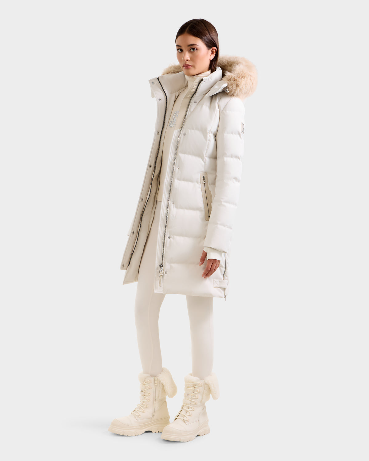 A woman wears the Rudsak KEHLANI H - 8125643 Vanilla long white down parka with a fur-lined hood, paired with white pants and winter boots, standing slightly to the side against a plain light background.