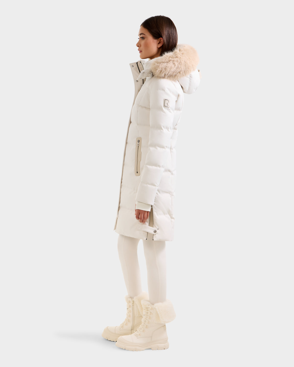 A woman stands in profile wearing the Rudsak KEHLANI H - 8125643 Vanilla, an off-white knee-length down winter coat with a fur-trimmed hood, matching leggings, and fur-lined boots against a plain light background.