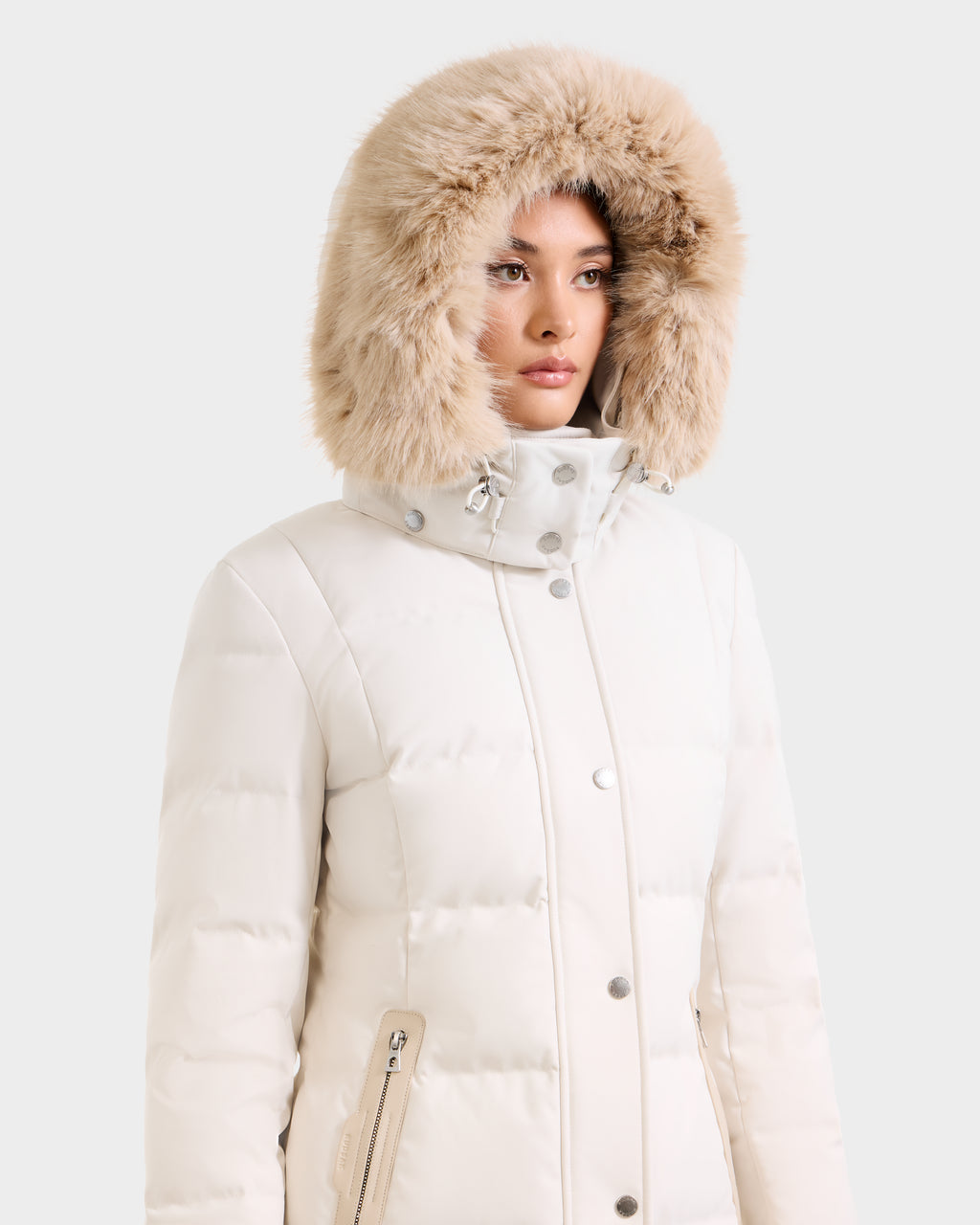 Wearing the Rudsak KEHLANI H - 8125643 Vanilla women's down parka with a fluffy fur-trimmed hood, a person looks to the side. The RDS-certified down provides warmth and ethical comfort against a plain backdrop.