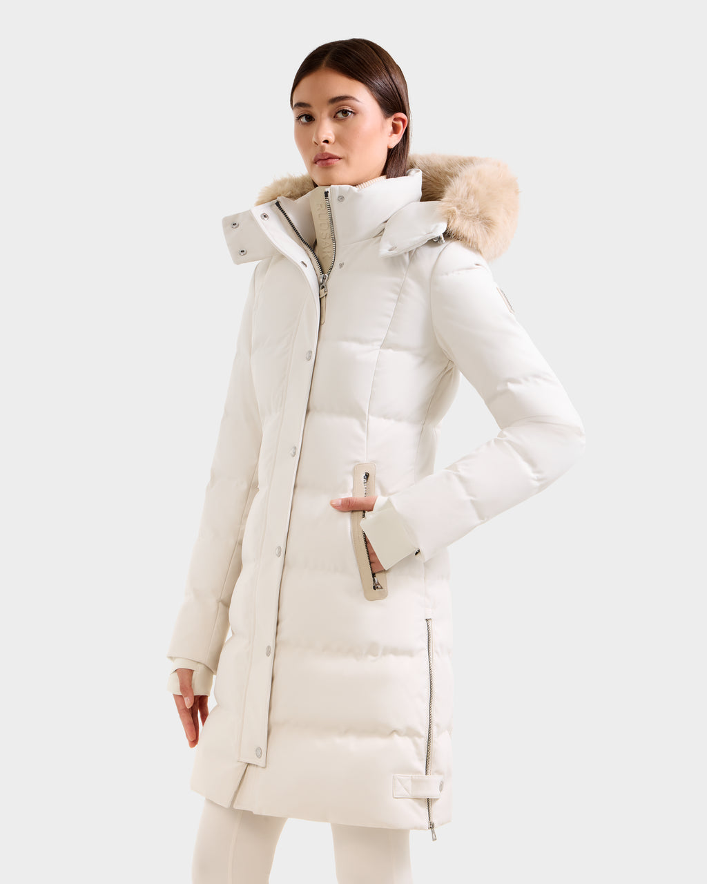 A woman stands slightly to the side against a plain background, wearing the Rudsak KEHLANI H - 8125643 Vanilla long white down parka with a fur-lined hood, zippered cuffs, side pockets, and RDS-certified down for warmth.