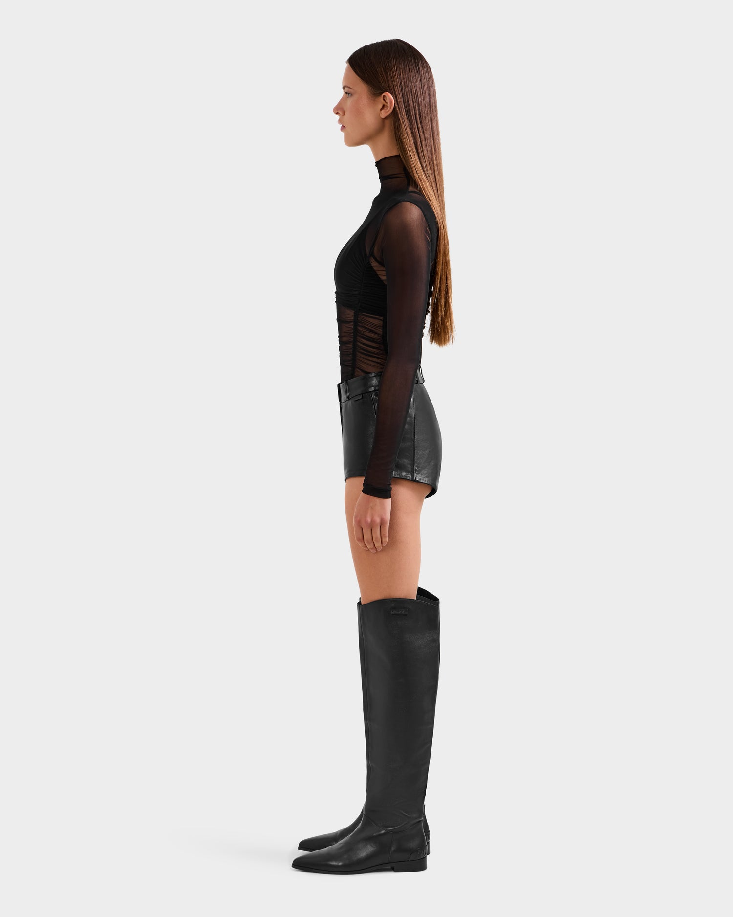 A woman with long brown hair stands in profile wearing Rudsaks KENNEDY - 8525610 Black mesh long-sleeve top, paired with high-rise shorts and knee-high black boots, against a plain white background.