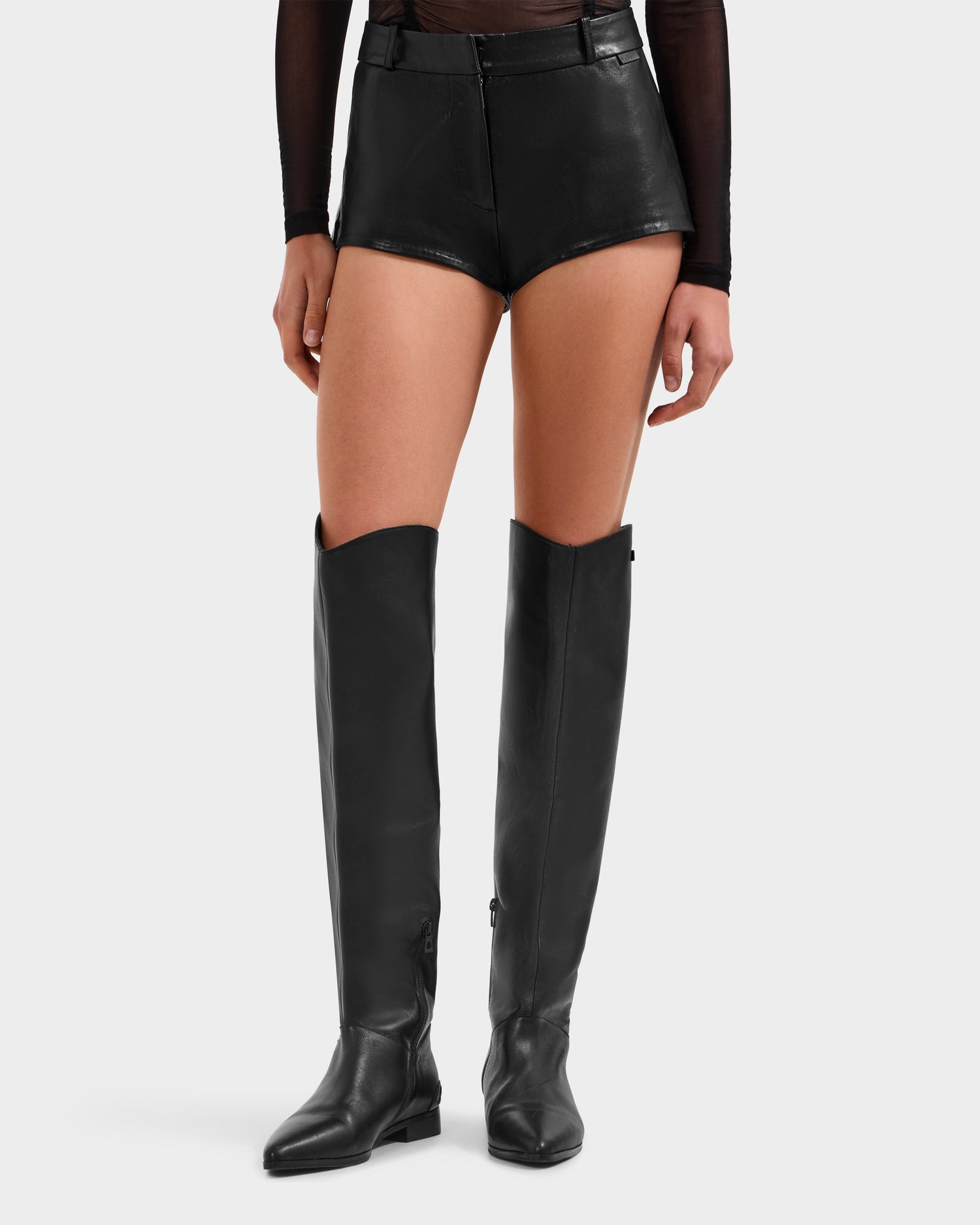 A person models the KENNEDY - 8525610 Black hot shorts by Rudsak with a sheer black top and knee-high black boots, posed against a plain light background.