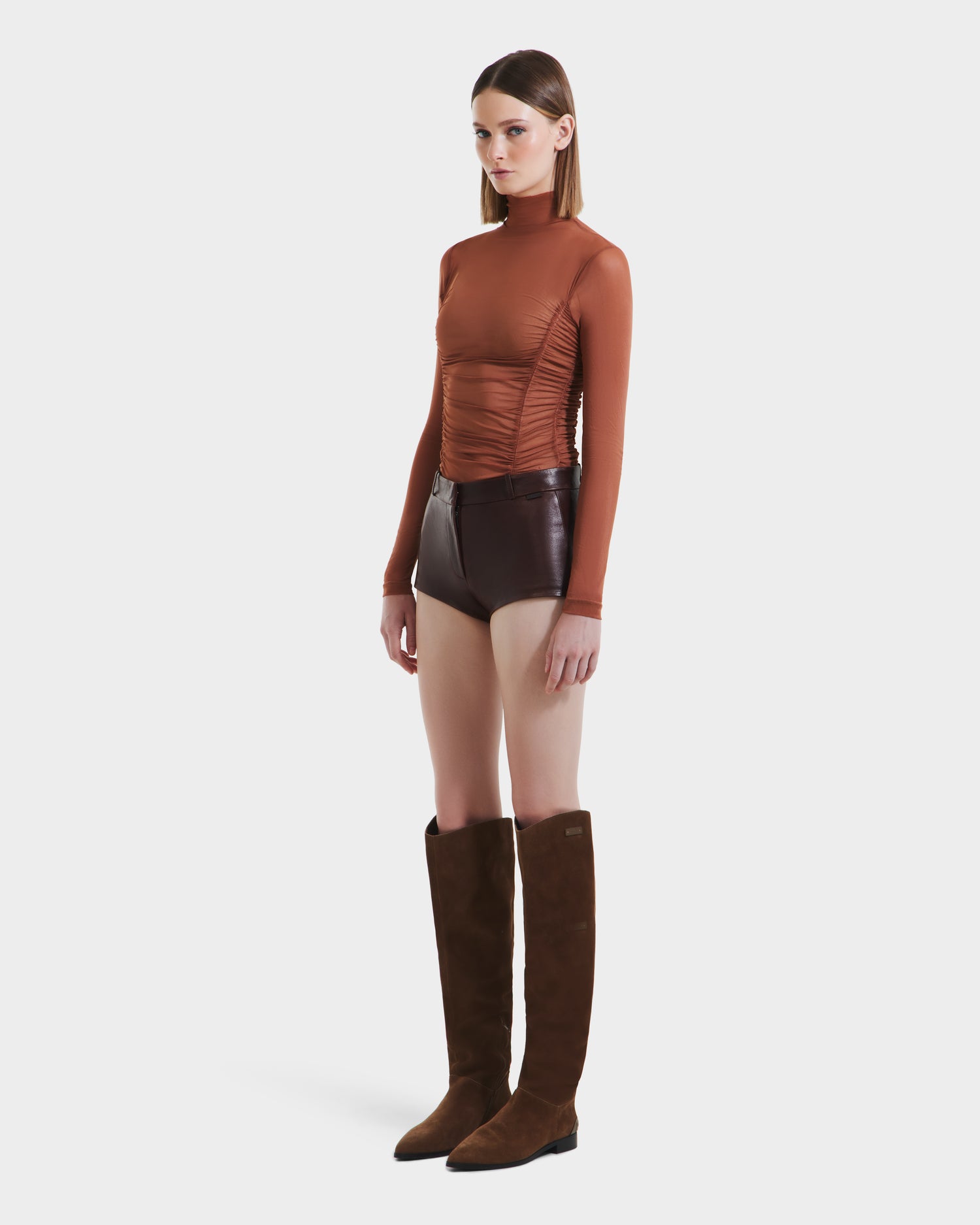 A woman wears the Rudsak KENNEDY - 8525610 Espresso fitted brown long-sleeve top, high-rise leather shorts, and knee-high brown suede boots, standing against a plain white background.