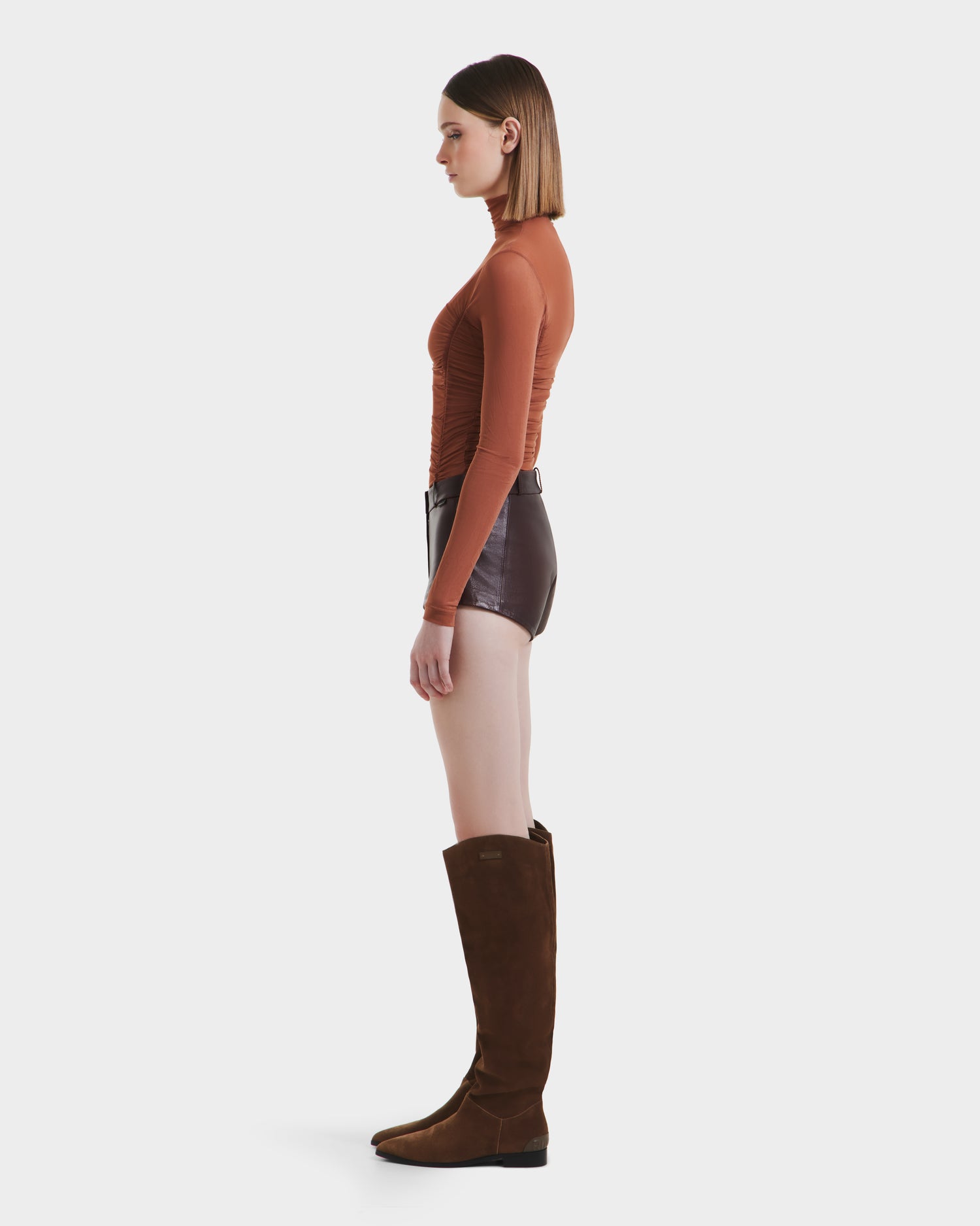 A woman stands in profile against a white background, wearing the Rudsak KENNEDY - 8525610 Espresso knee-high suede boots, styled with a long-sleeve rust top and high-rise leather shorts.