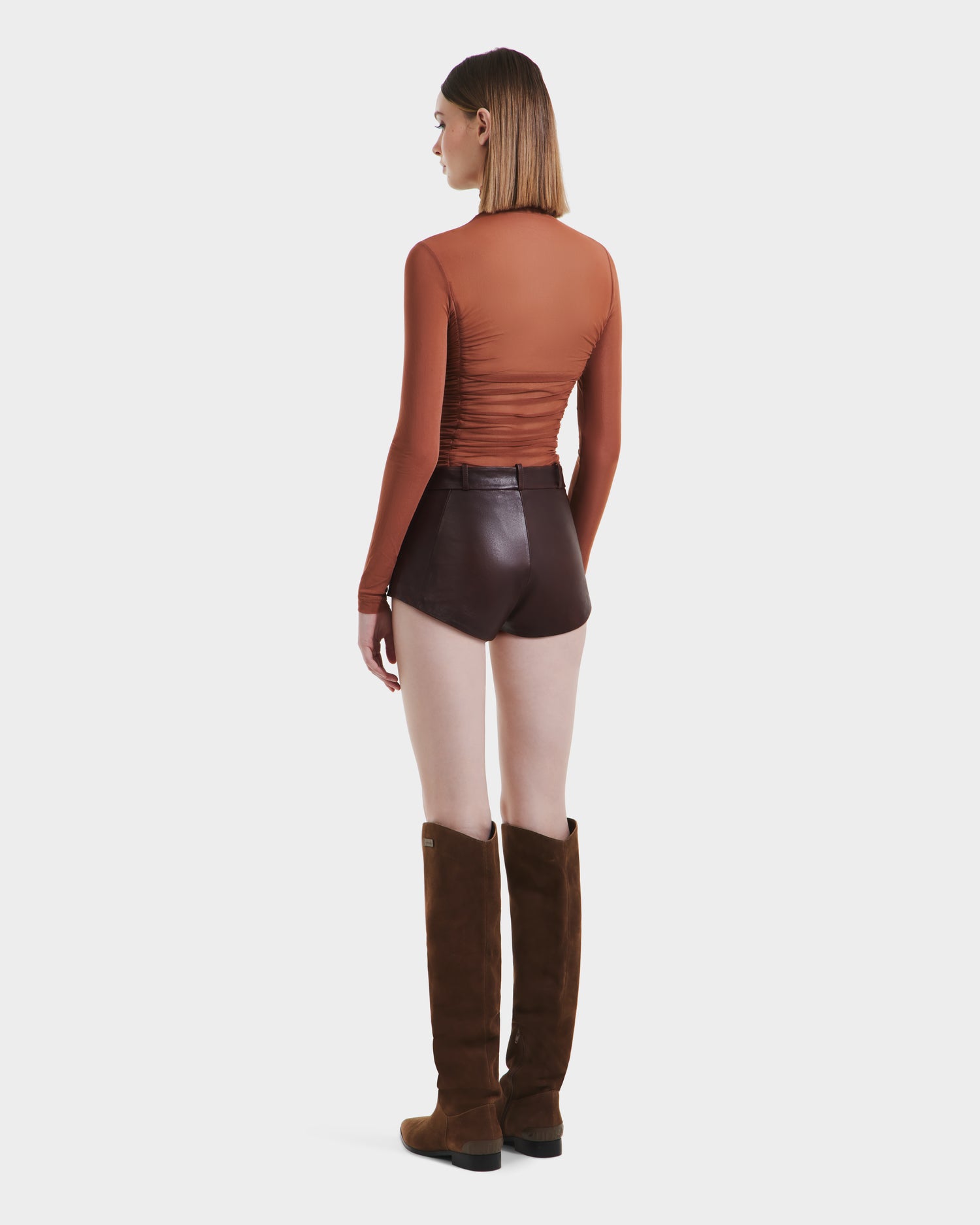 A woman faces away, wearing Rudsaks KENNEDY - 8525610 Espresso long-sleeve top, high-rise leather shorts, and knee-high brown suede boots against a plain white background.