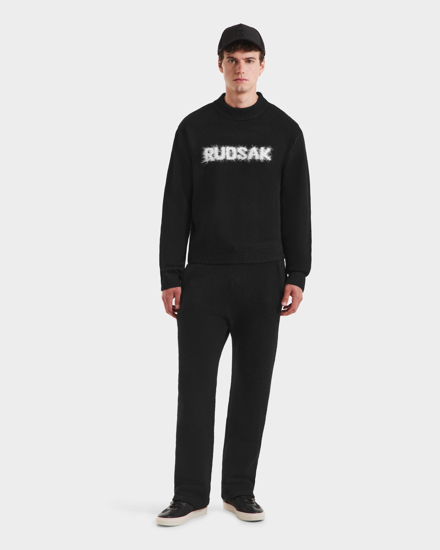 A man stands against a white background wearing the Rudsak KINLEY - 9525555 Black M unisex textured knit crewneck sweater with a bold logo, paired with black pants, sneakers, and a cap.