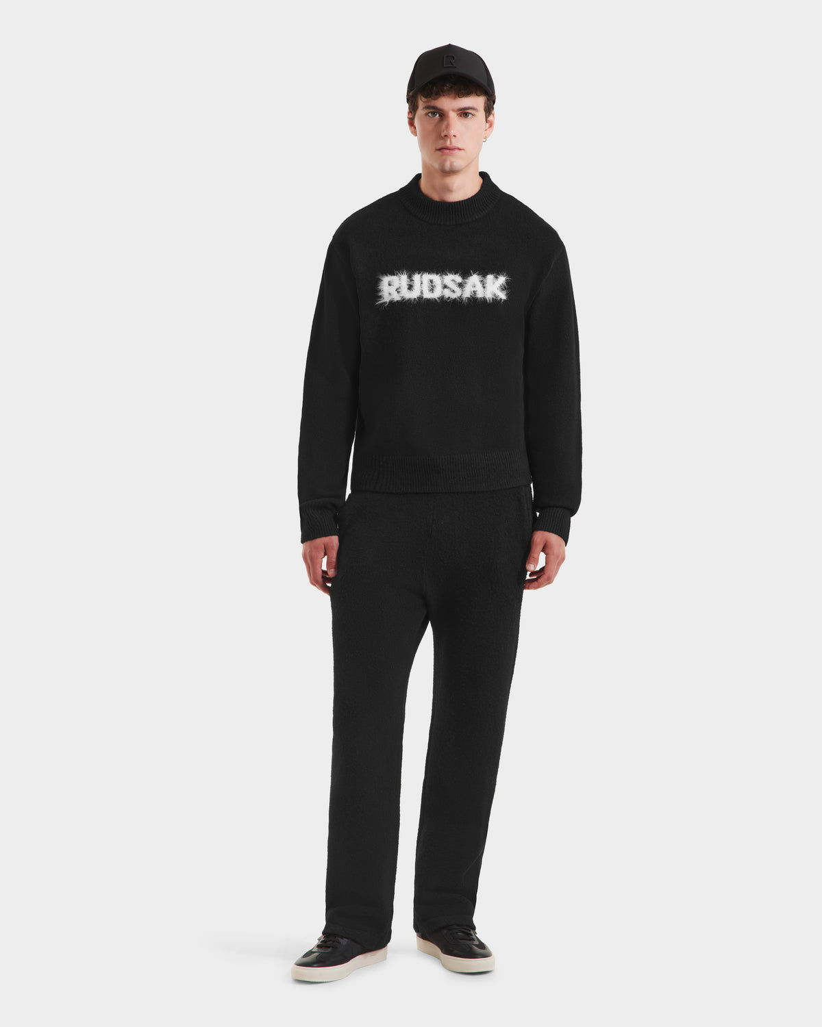 A man stands against a white background wearing the Rudsak KINLEY - 9525555 Black M unisex textured knit crewneck sweater with a bold logo, paired with black pants, sneakers, and a cap.