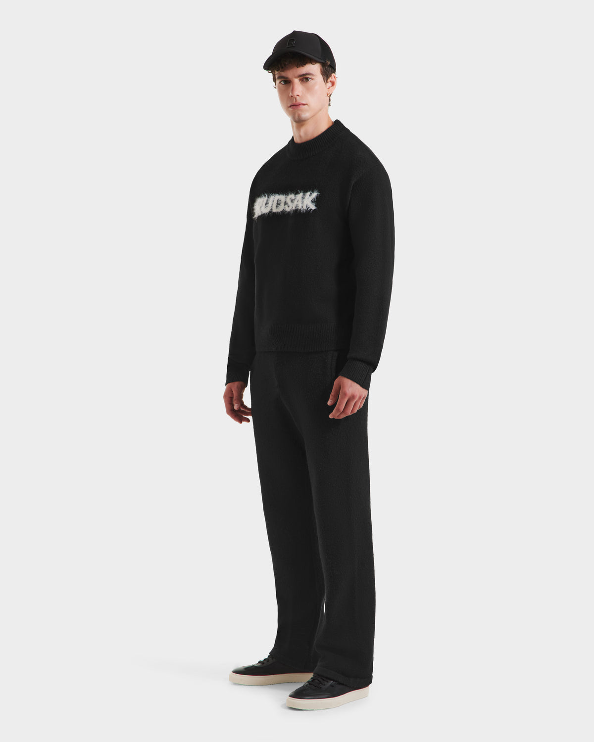 A person stands against a white background wearing the Rudsak KINLEY - 9525555 Black M crewneck sweater, paired with a black baseball cap, black pants, and black-and-white sneakers.