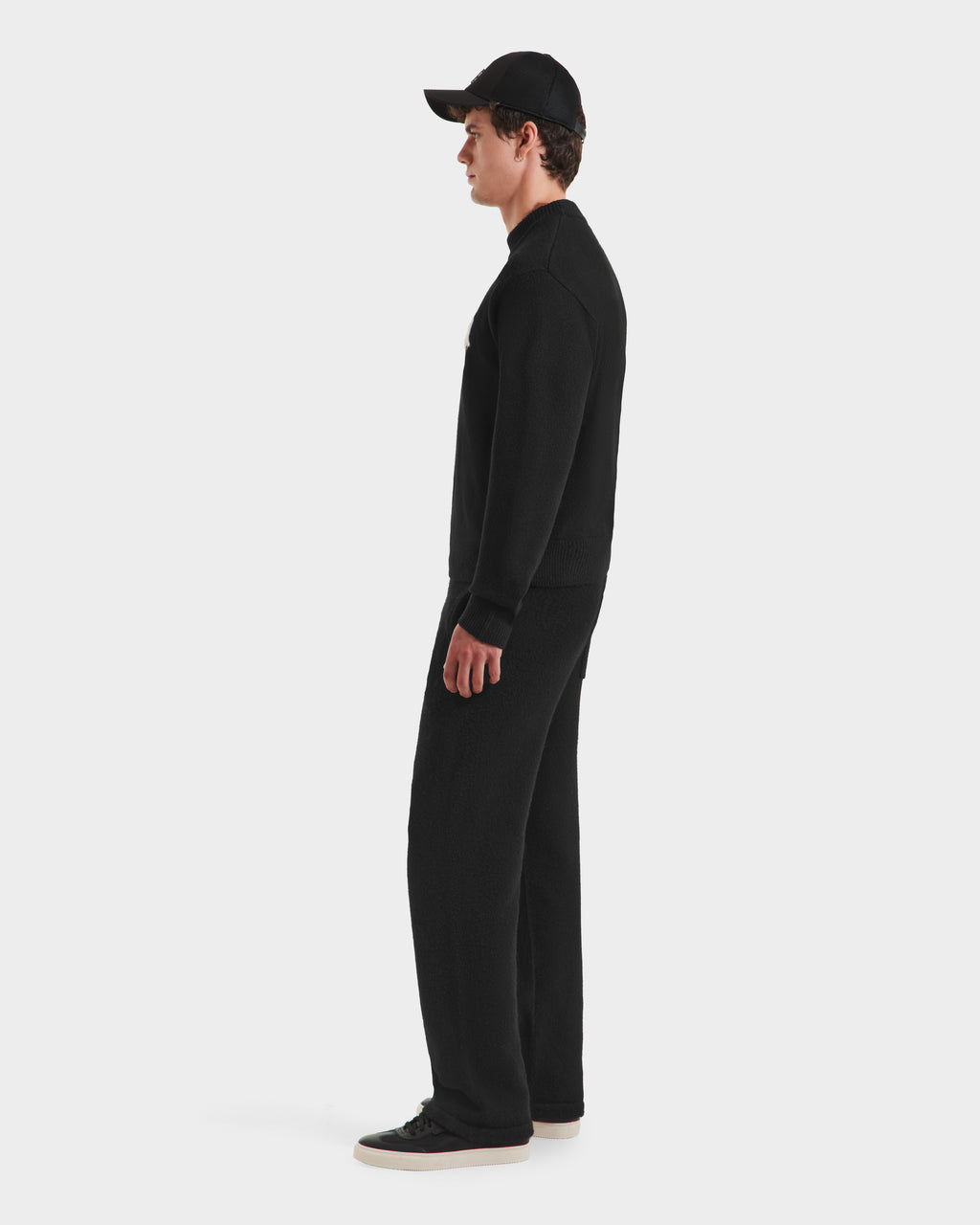 A person in profile wears a black Rudsak KINLEY - 9525555 unisex crewneck sweater, black pants, and black sneakers with white soles against a plain white background.