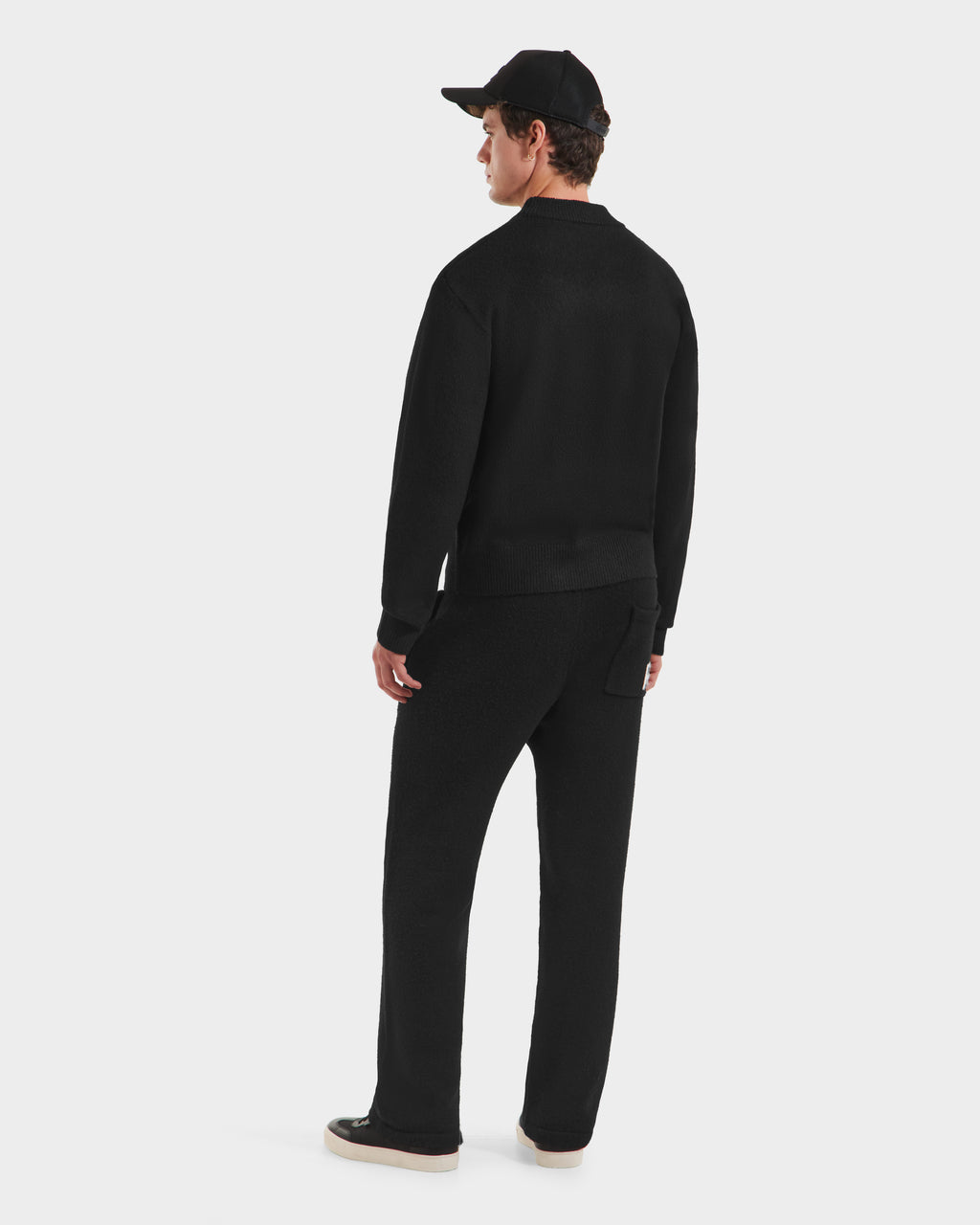 A person wearing the Rudsak KINLEY - 9525555 Black M unisex crewneck sweater with a subtle logo, plus black pants, cap, and shoes stands facing away against a plain white background.