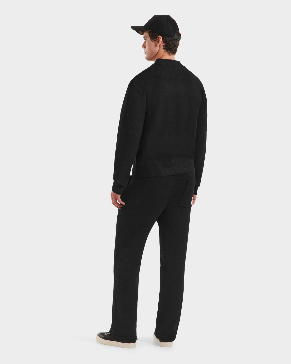 A person wearing the Rudsak KINLEY - 9525555 Black M unisex crewneck sweater with a subtle logo, plus black pants, cap, and shoes stands facing away against a plain white background.