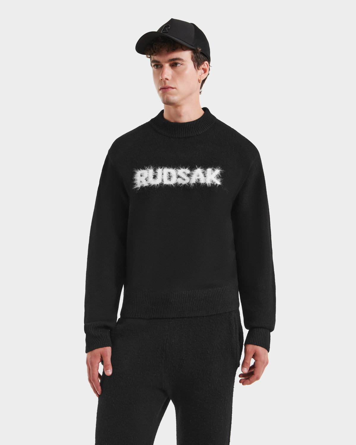 A young man in the Rudsak KINLEY - 9525555 Black M sweater, a unisex crewneck design, pairs it with black pants and stands against a plain white background.