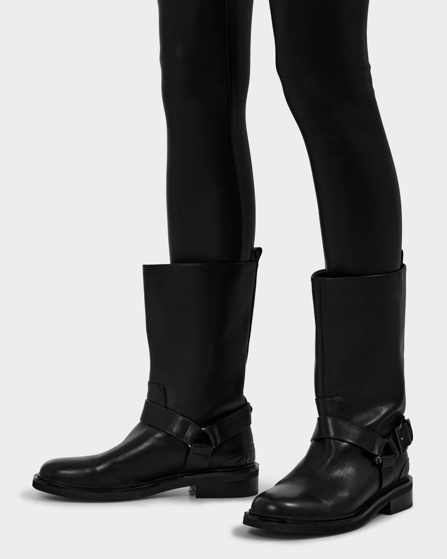 A person wearing the Rudsak KIRRA - 8225558 Black knee-high leather moto boots with harness straps and a slight heel over slim black pants, posed against a plain light background.