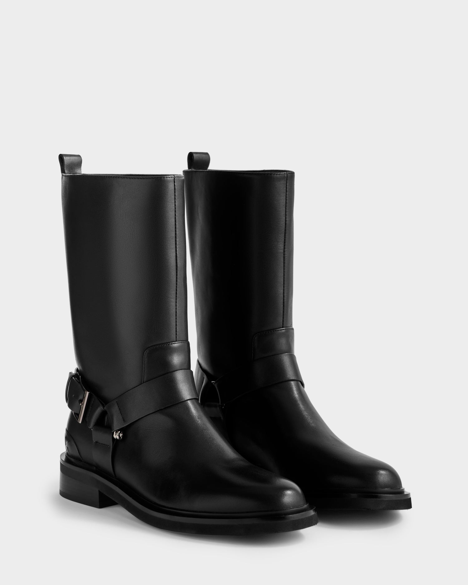 The Rudsak KIRRA - 8225558 Black are women’s black leather moto boots with low heels, harness straps with metal buckles, and pull tabs—fashionably displayed on a plain white background.