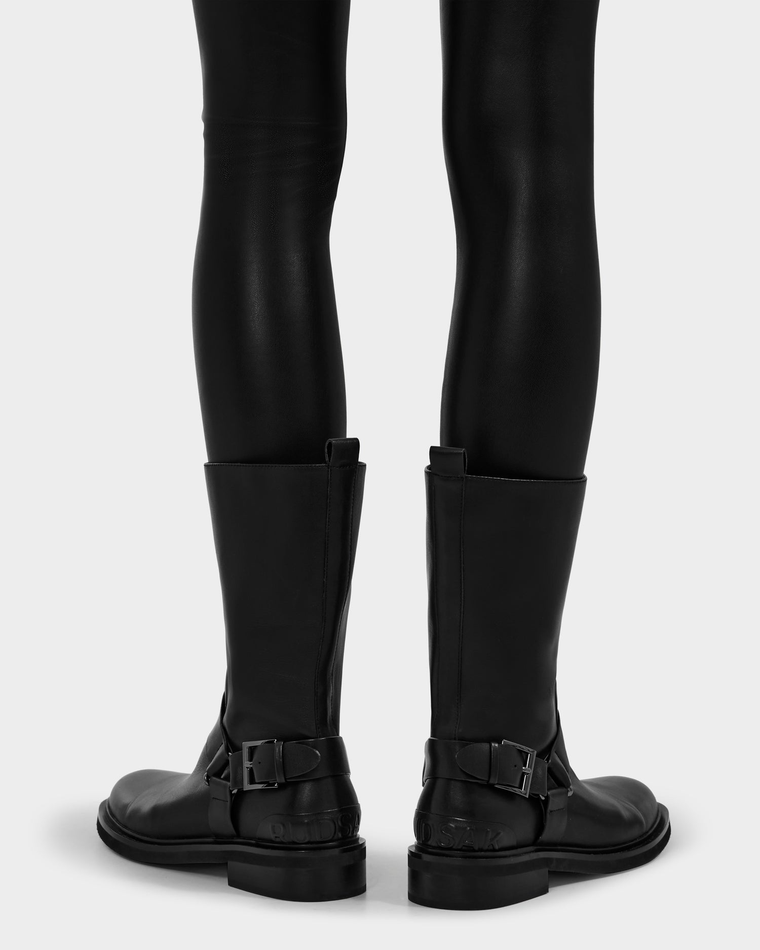 A person wearing Rudsak KIRRA - 8225558 Black leather pants and tall moto boots with buckles, shown from the back against a plain light background.