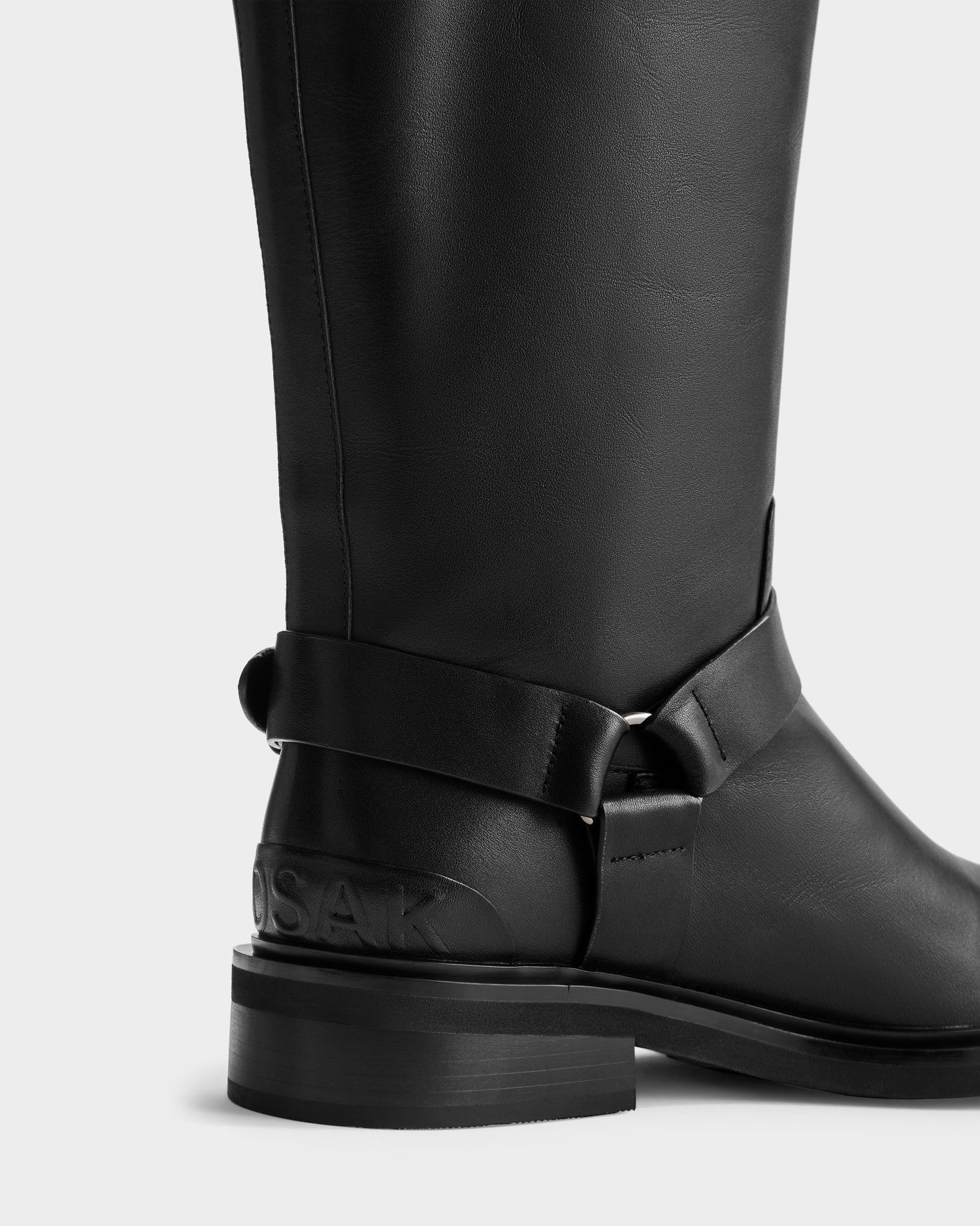 A close-up of the Rudsak KIRRA - 8225558 Black leather knee-high moto boot with a thick heel, ankle strap detail, and embossed heel text, set against a white background—ideal for elevating any women's fashion boots collection.