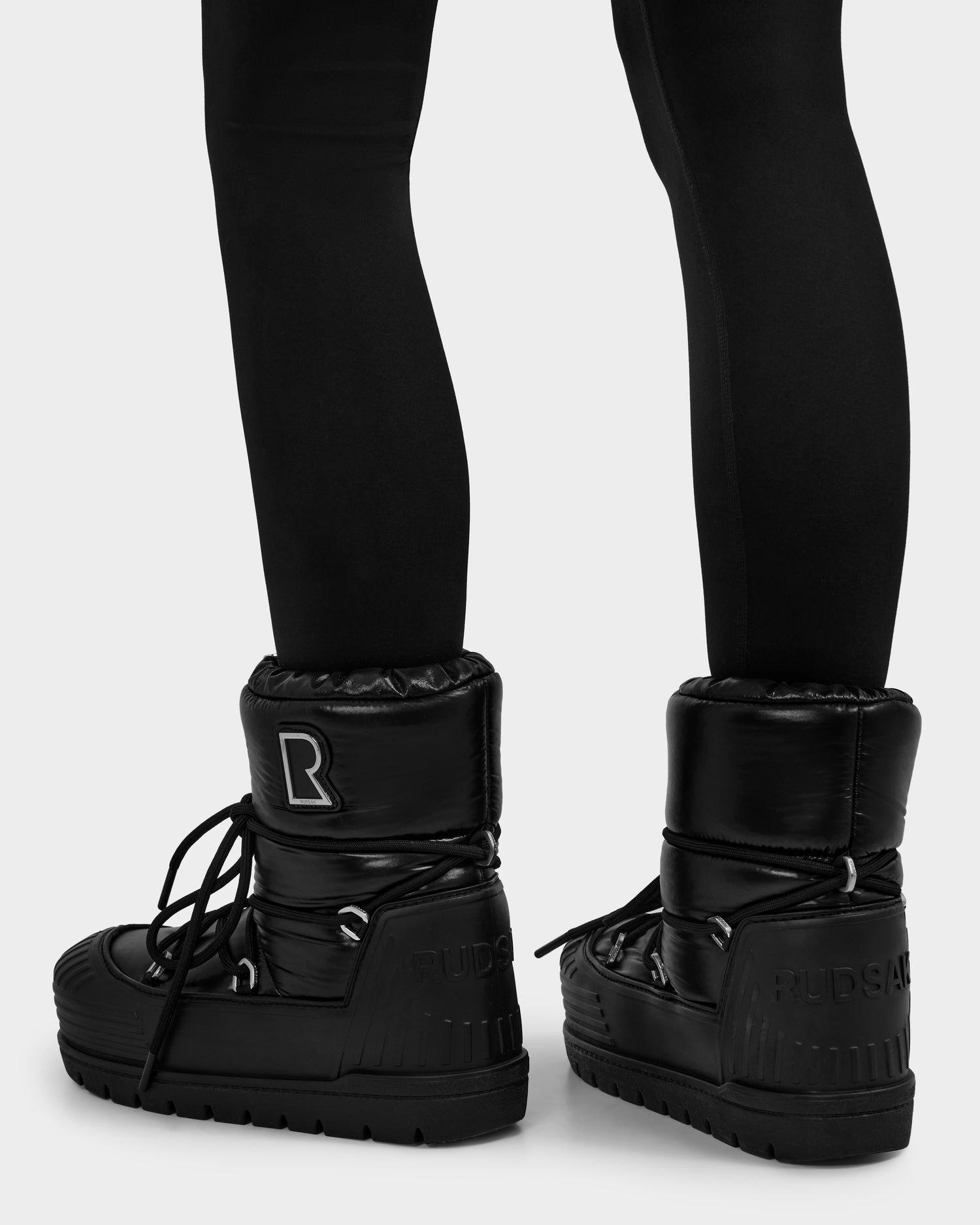 A person is shown from the back in black leggings and Rudsak's KIKA - L224506 Black women’s winter boots, featuring a shiny finish, chunky rubber sole, and laces, set against a plain light background—perfect for winter outings.