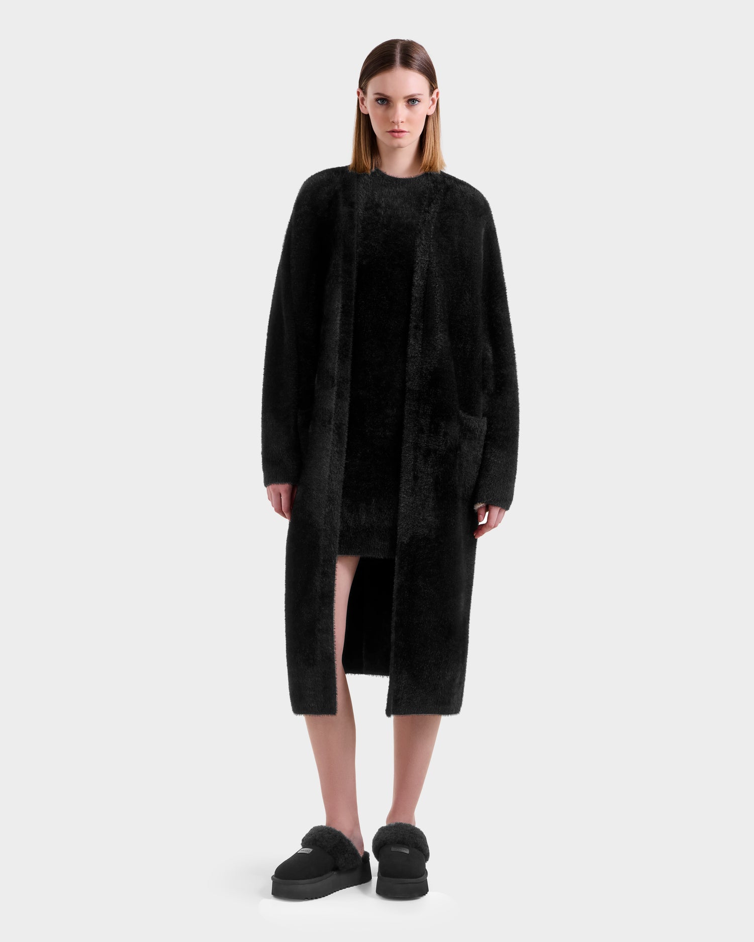 A woman faces forward in a Rudsak LACEY - 8525694 Black fuzzy coat, layered over a black dress with matching fuzzy slippers, set against a plain light gray background.