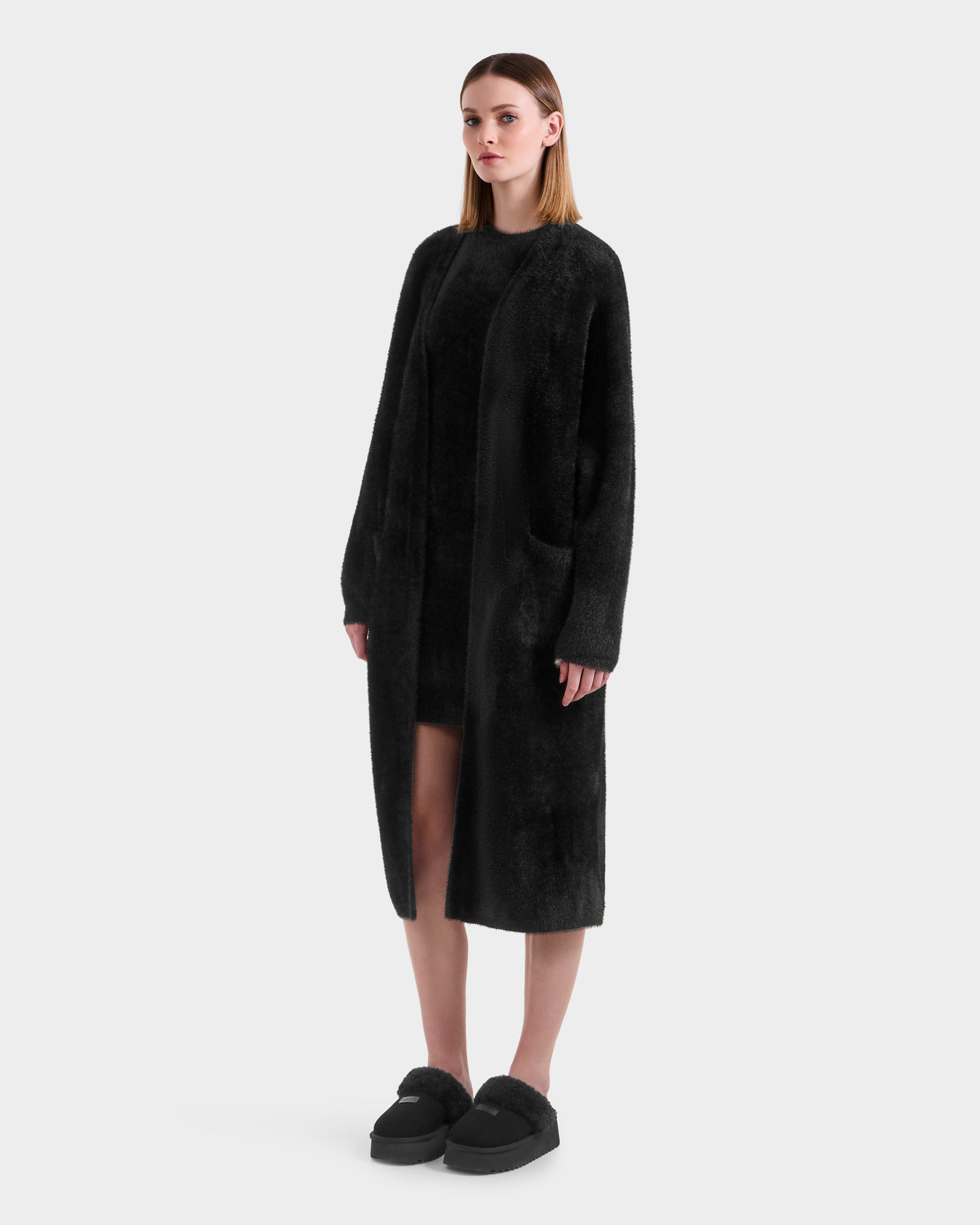 A woman faces forward wearing the Rudsak LACEY - 8525694 Black fuzzy sweater coat over a matching dress and slip-on shoes, perfect for cozy layering, set against a plain light gray background.