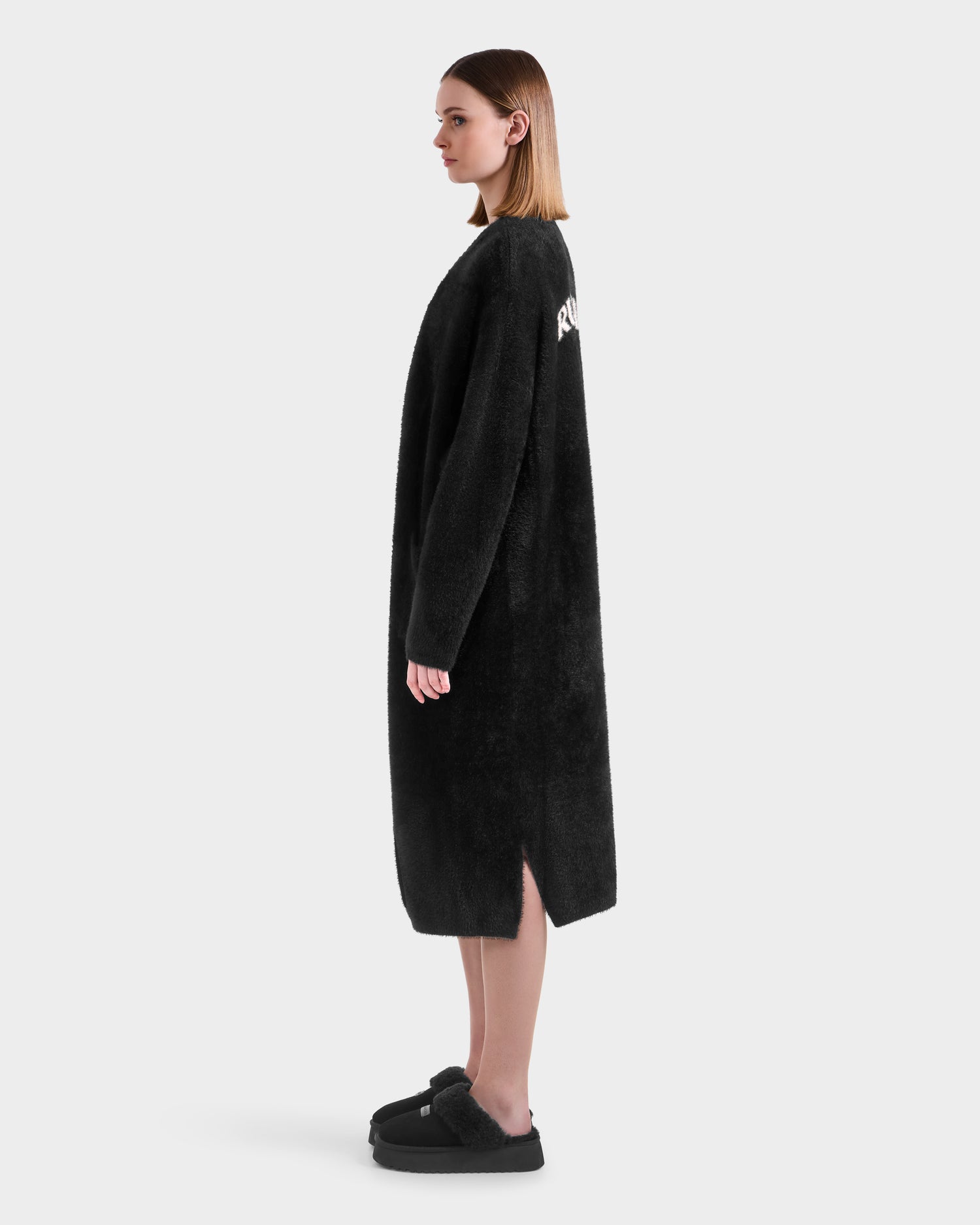 A woman in profile wears the Rudsak LACEY - 8525694 Black long fuzzy sweater coat with side slits, styled with matching black slippers, set against a plain white background.