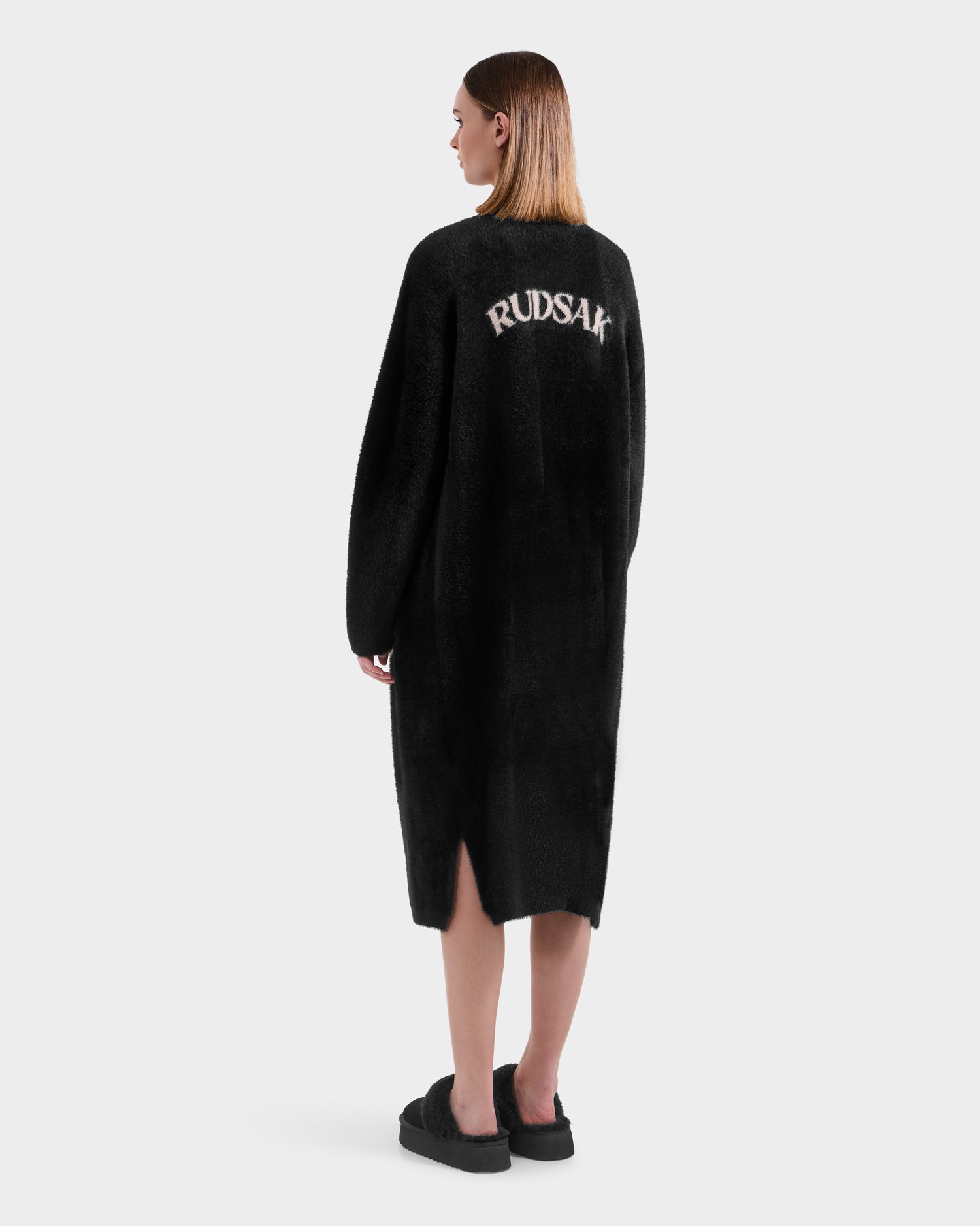 A person with straight, shoulder-length hair wears the Rudsak LACEY - 8525694 Black fuzzy sweater coat, featuring "RUDSAK" in white on the back. They’re turned away against a plain light background—perfect for cozy layering.