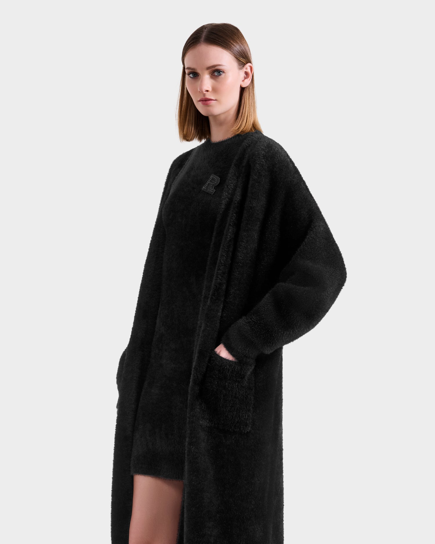 A woman with straight, shoulder-length hair wears the Rudsak LACEY - 8525694 Black long textured coat with large sleeves and pockets over a black outfit, standing against a plain light gray background.