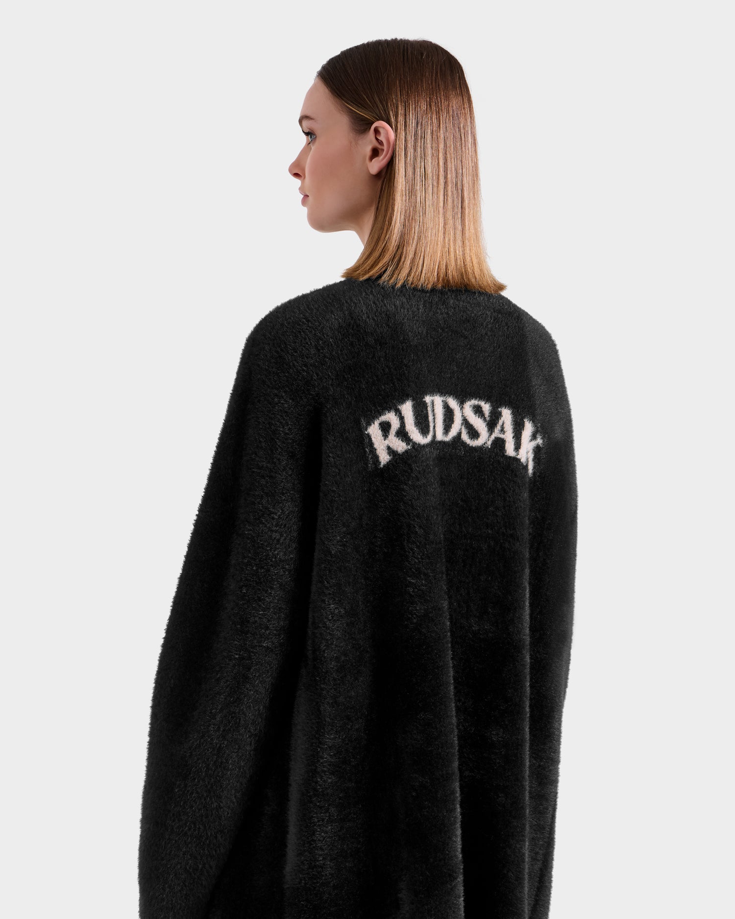 A woman with straight, shoulder-length hair models the Rudsak LACEY - 8525694 Black fuzzy coat, featuring bold white "RUDSAK" lettering on the back. She faces away to showcase the design against a plain white background.