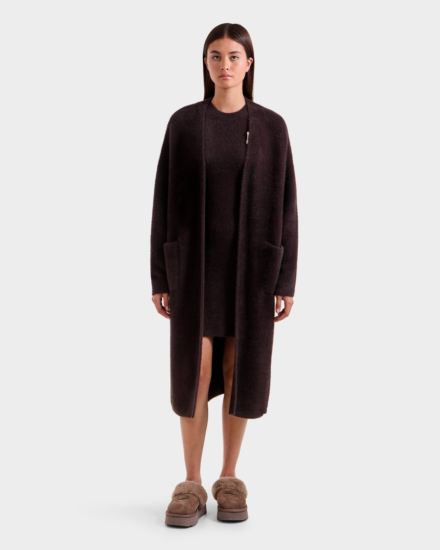 A woman stands facing forward in the Rudsak LACEY - 8525694 Espresso, a cozy dark brown fuzzy cardigan, layered over a matching dress with brown fuzzy slippers, set against a plain light background.