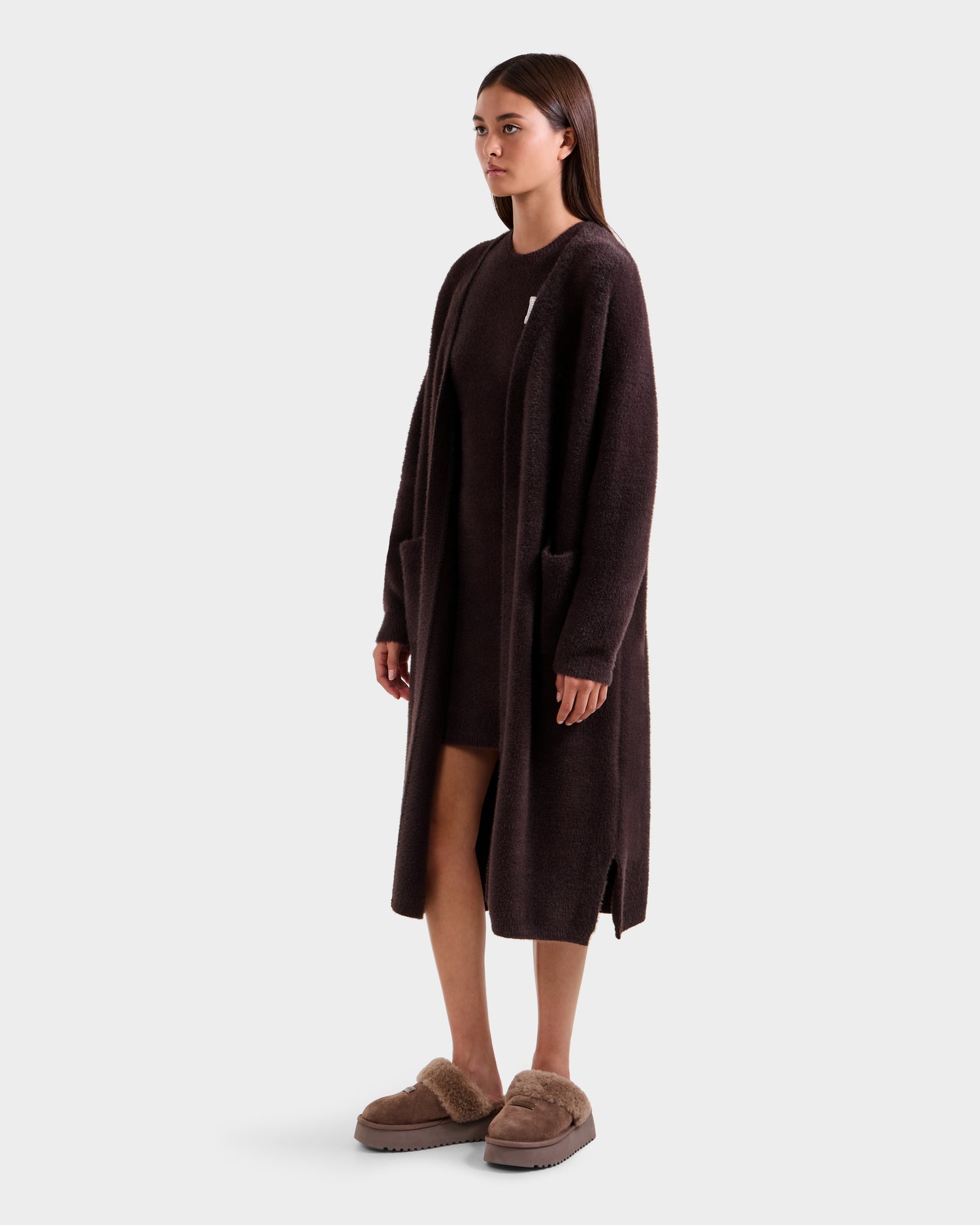 A woman poses in the LACEY - 8525694 Espresso by Rudsak, layering a long cardigan over a matching dress with beige fuzzy slippers against a plain light gray background, gazing slightly to the side.