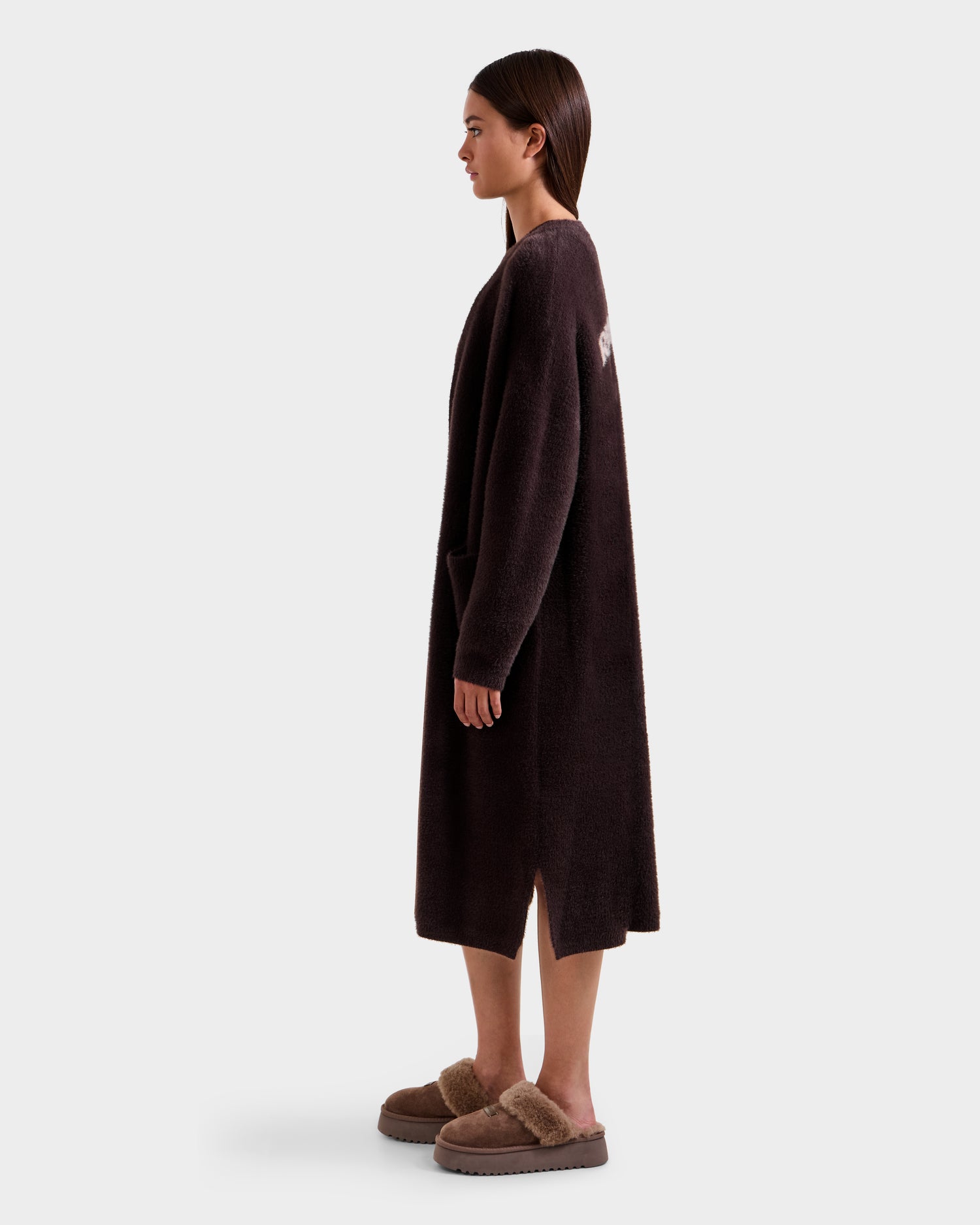 A woman in profile models the Rudsak LACEY - 8525694 Espresso, a long dark brown fuzzy cardigan, paired with matching fur-trimmed slippers. She faces right against a plain light background, highlighting cozy layering style.