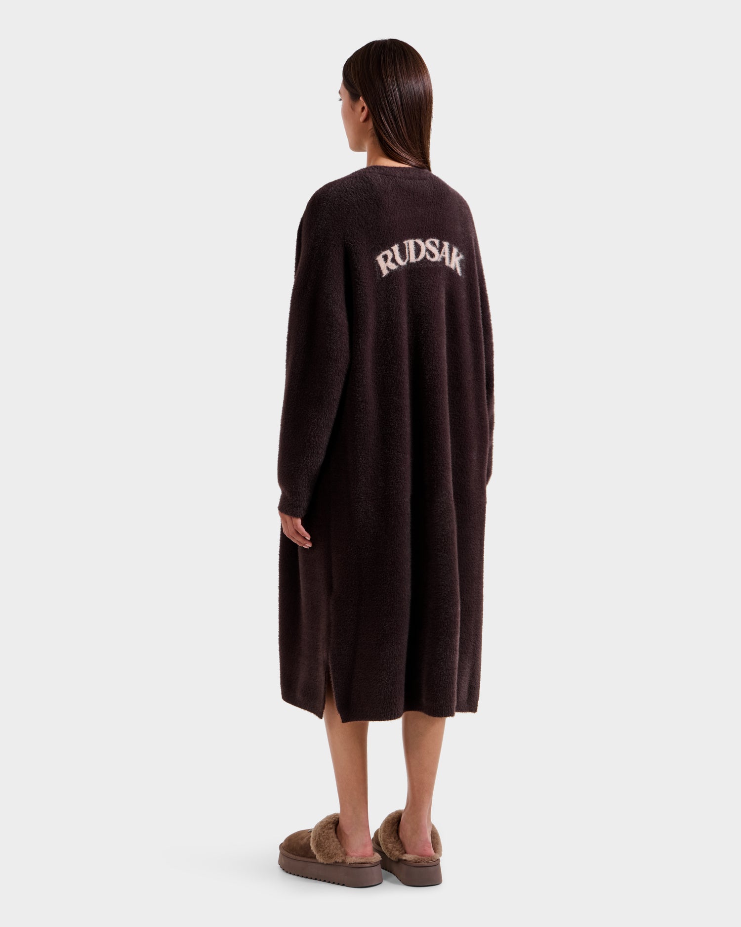 A woman faces away, wearing the Rudsak LACEY - 8525694 Espresso fuzzy cardigan with bold white RUDSAK lettering on the back, paired with matching slippers against a plain light grey background.