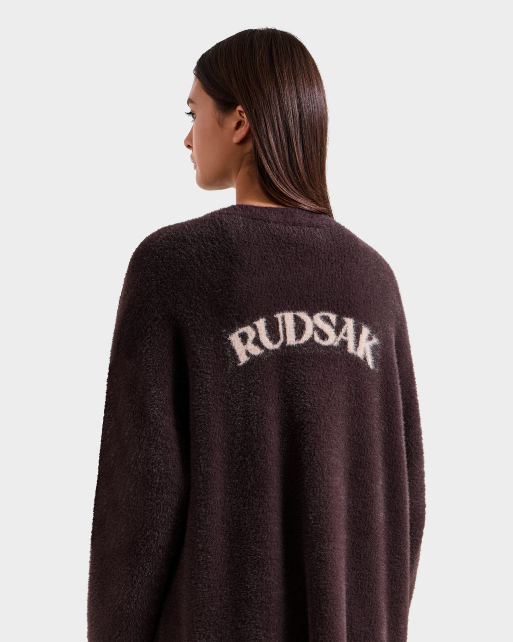 A woman with straight brown hair faces away, wearing Rudsak’s LACEY - 8525694 Espresso dark fuzzy coat with “RUDSAK” in bold white letters across the back, set against a plain light background.