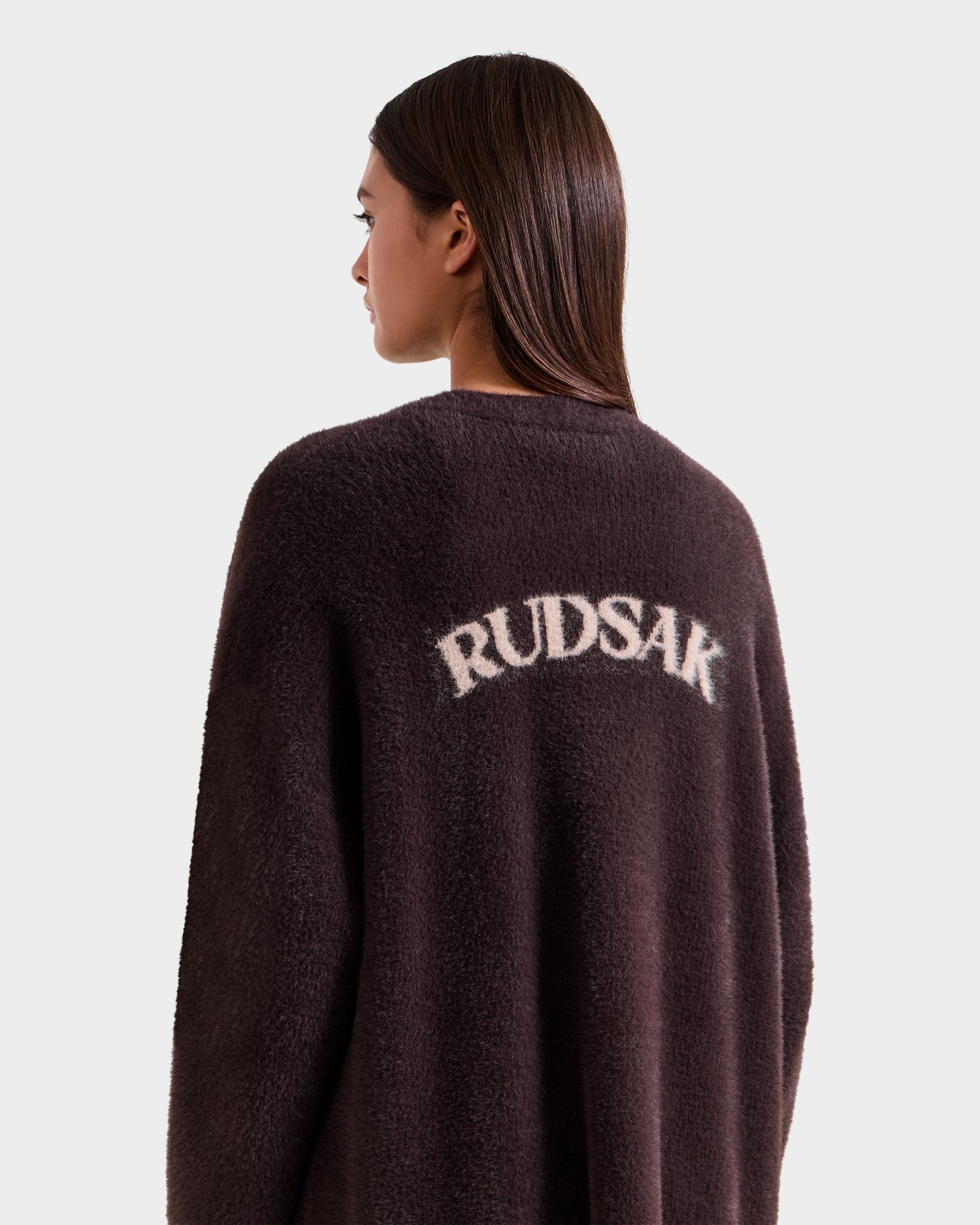 A woman with straight brown hair faces away, wearing Rudsak’s LACEY - 8525694 Espresso dark fuzzy coat with “RUDSAK” in bold white letters across the back, set against a plain light background.