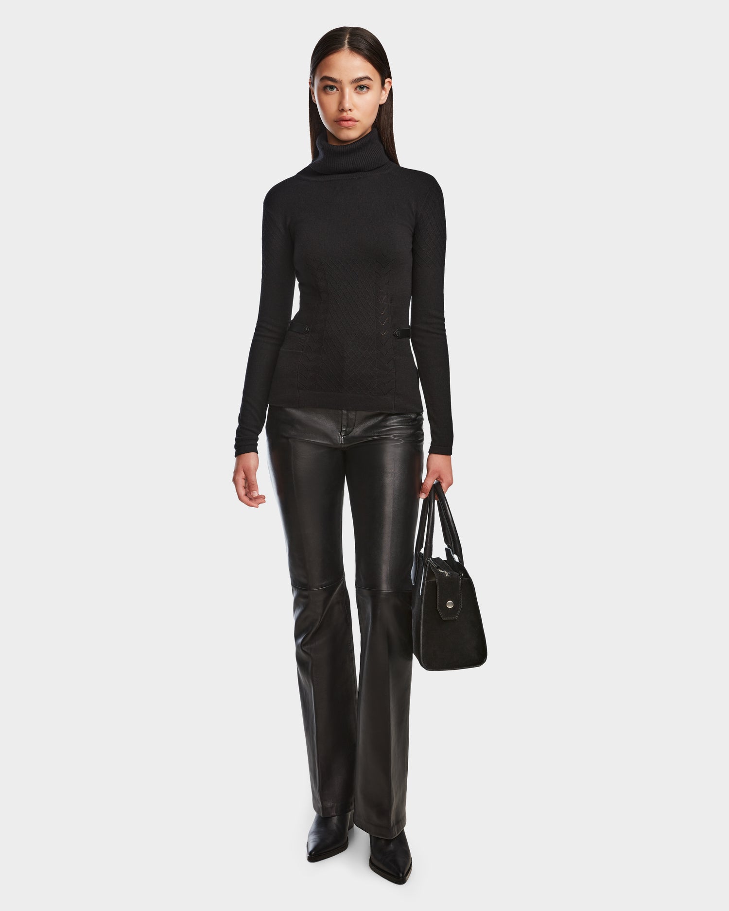 A woman with long, straight hair stands facing forward against a white background, wearing the Rudsak LAURA - 8524596 Black knit turtleneck sweater, black vegan leather-trimmed pants, pointed black shoes, and holding a black handbag.
