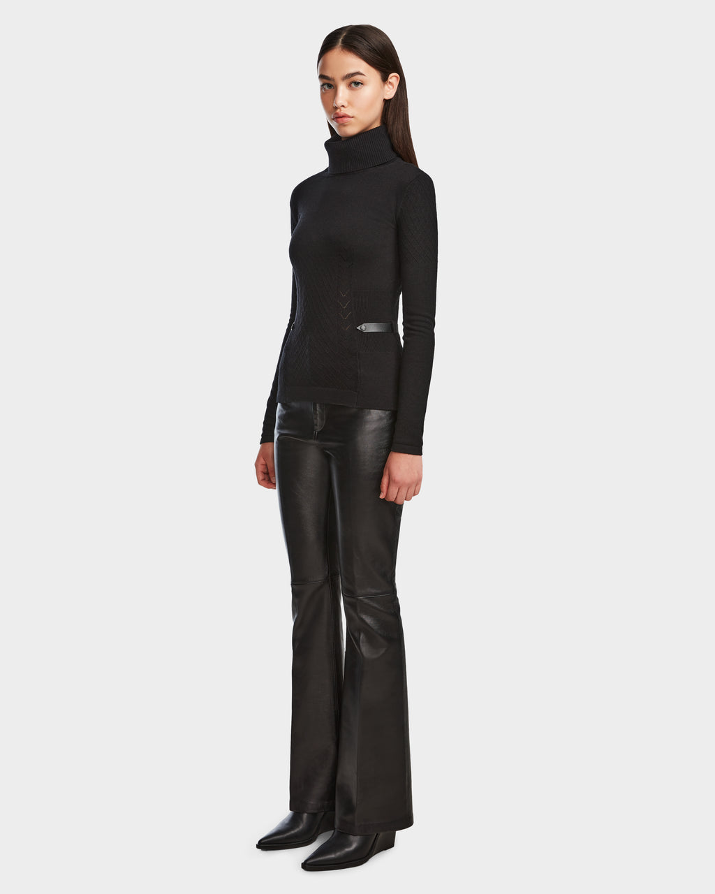A woman with straight brown hair and a neutral expression stands against a plain background wearing Rudsaks LAURA - 8524596 Black knit turtleneck sweater, black leather flared pants with vegan leather trim, and black boots.