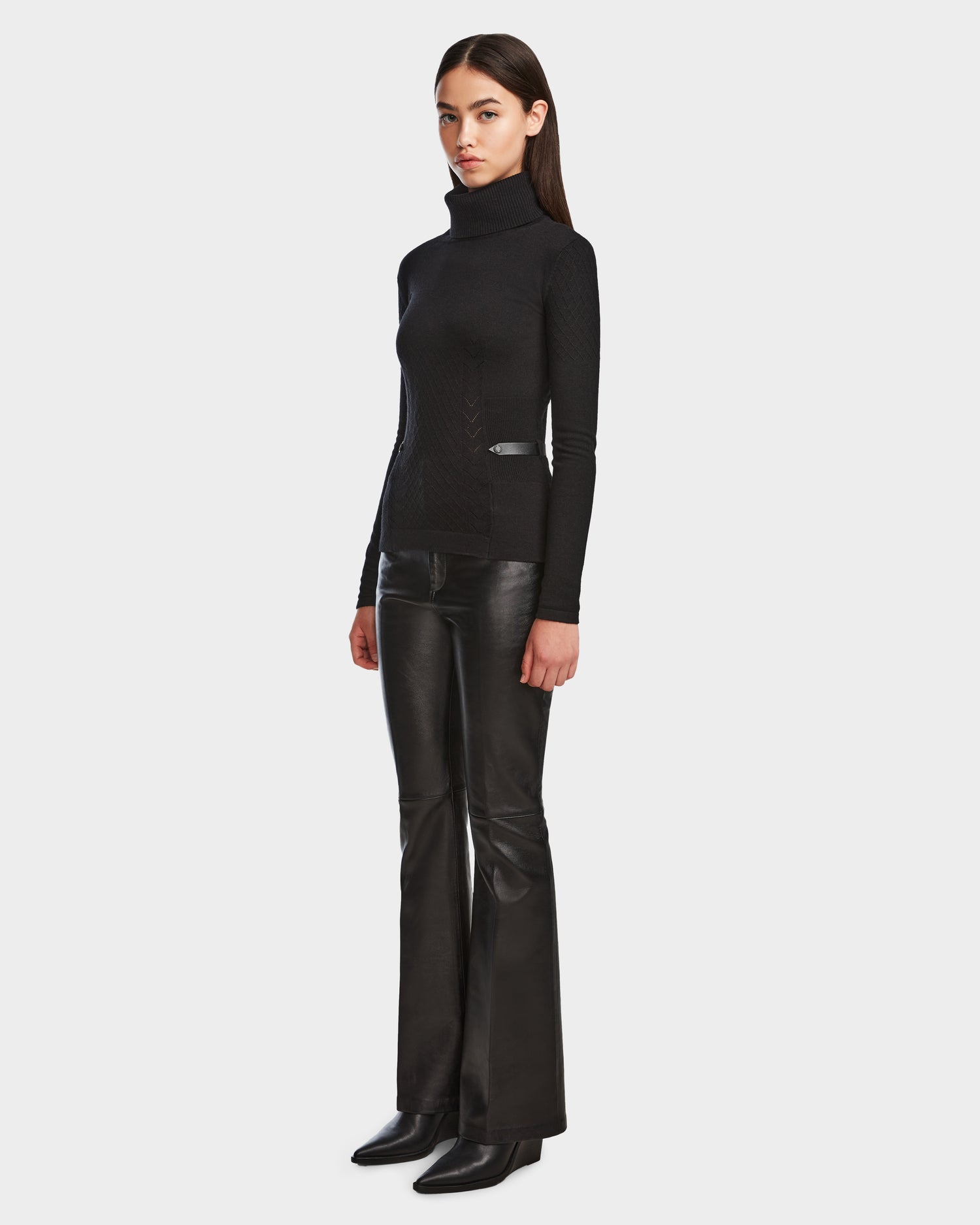 A woman with straight brown hair and a neutral expression stands against a plain background wearing Rudsaks LAURA - 8524596 Black knit turtleneck sweater, black leather flared pants with vegan leather trim, and black boots.