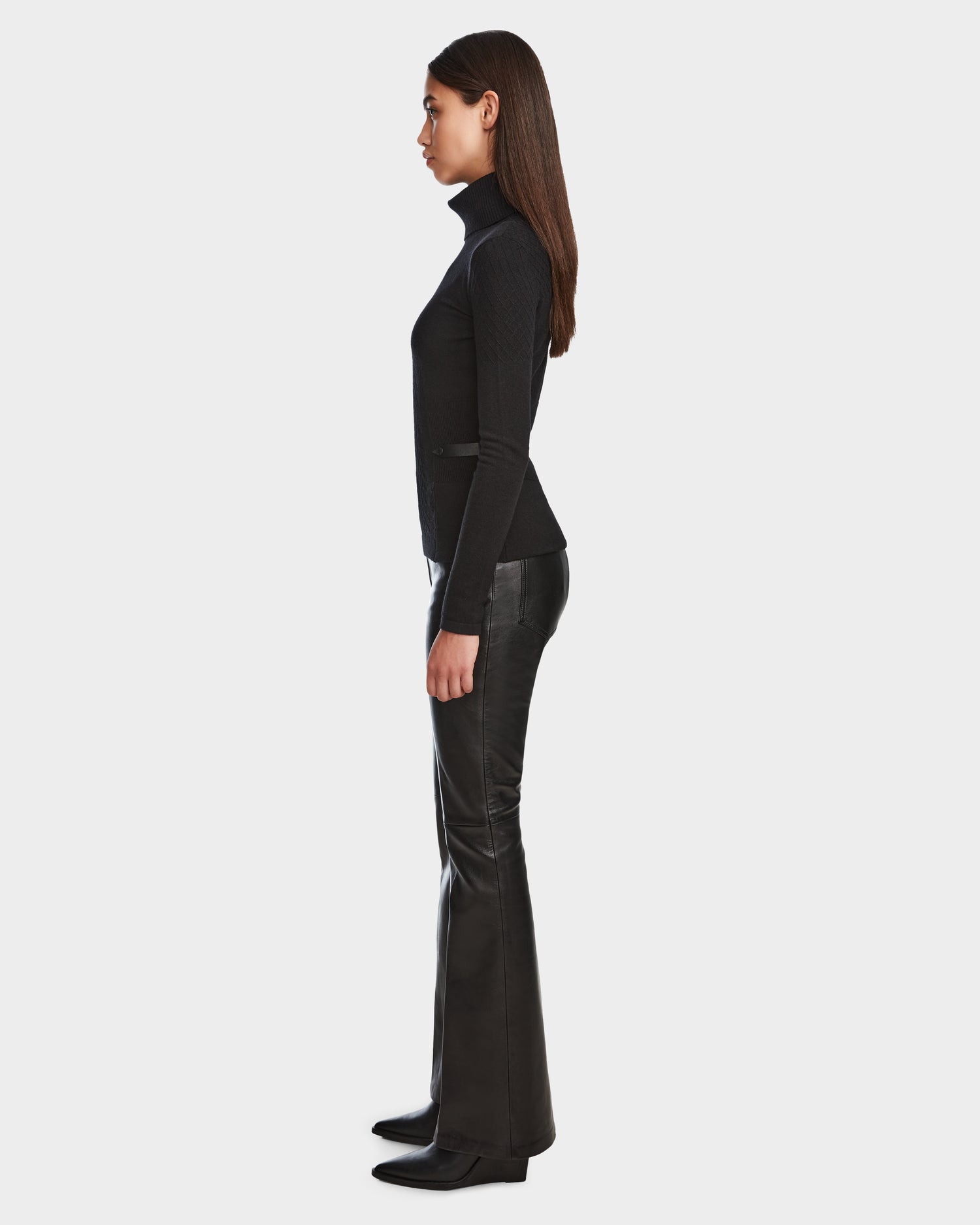 A woman with straight brown hair stands in profile, wearing the Rudsak LAURA - 8524596 Black womens knit turtleneck sweater and black boots, paired with black leather pants with vegan leather trim, against a plain white background.