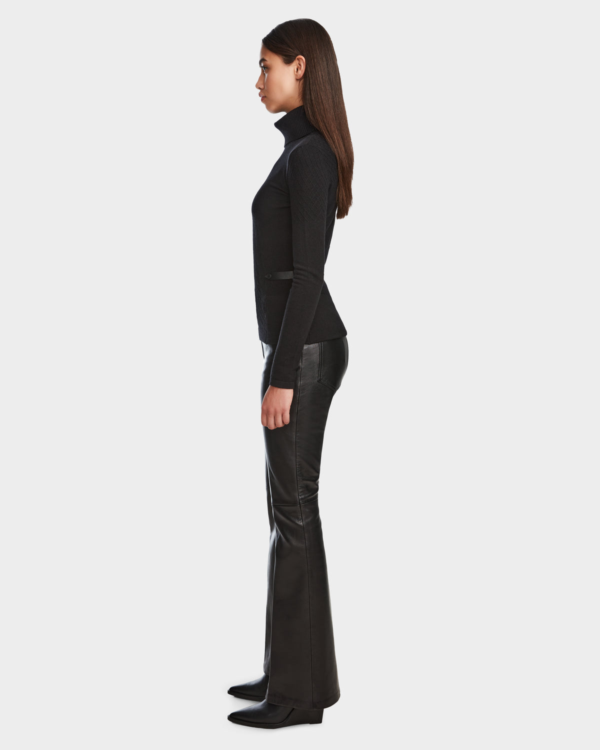 A woman with straight brown hair stands in profile, wearing the Rudsak LAURA - 8524596 Black womens knit turtleneck sweater and black boots, paired with black leather pants with vegan leather trim, against a plain white background.