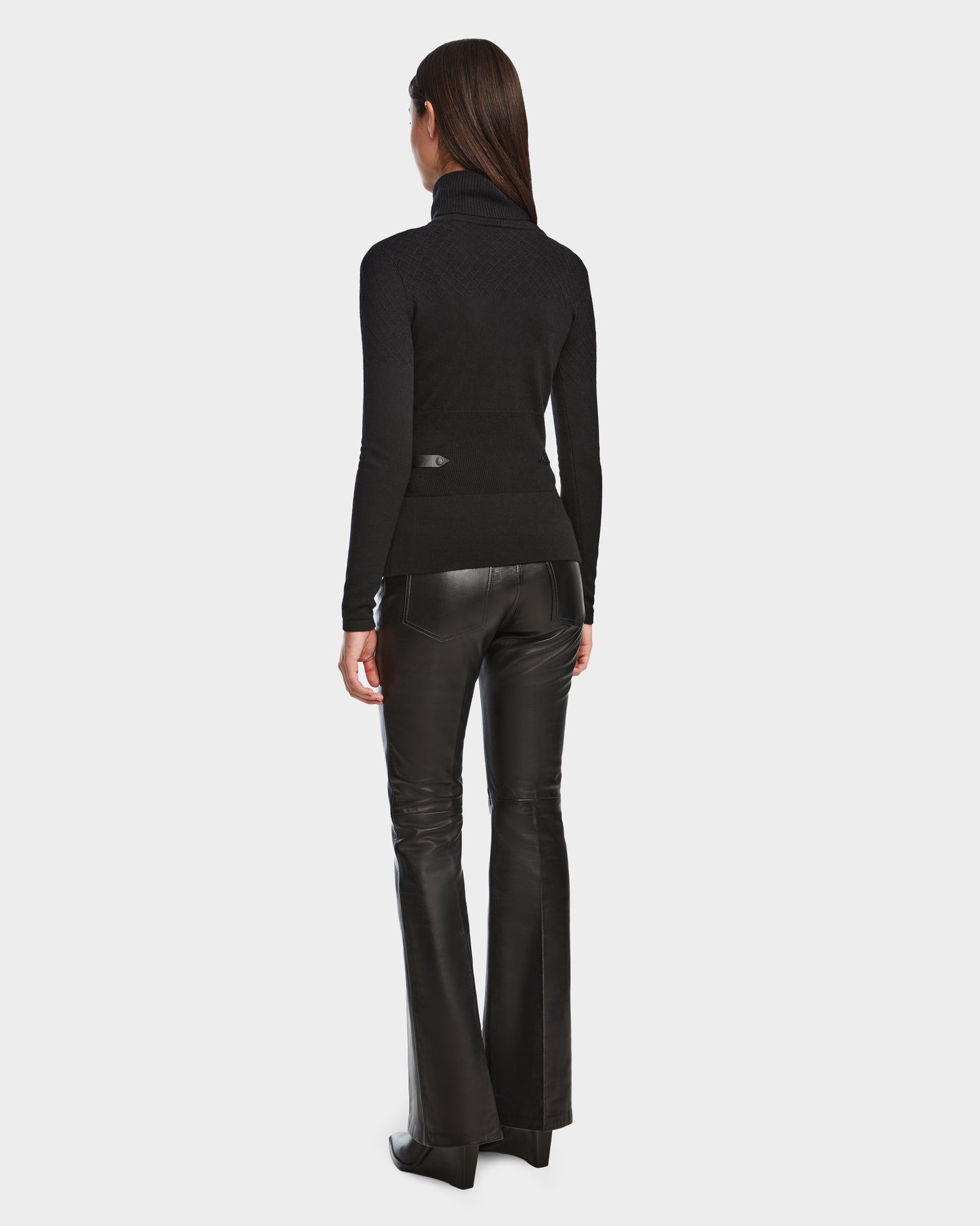 A woman with long brown hair faces away, wearing the Rudsak LAURA - 8524596 Black women’s knit turtleneck sweater and black flared pants with vegan leather trim, set against a plain white background.