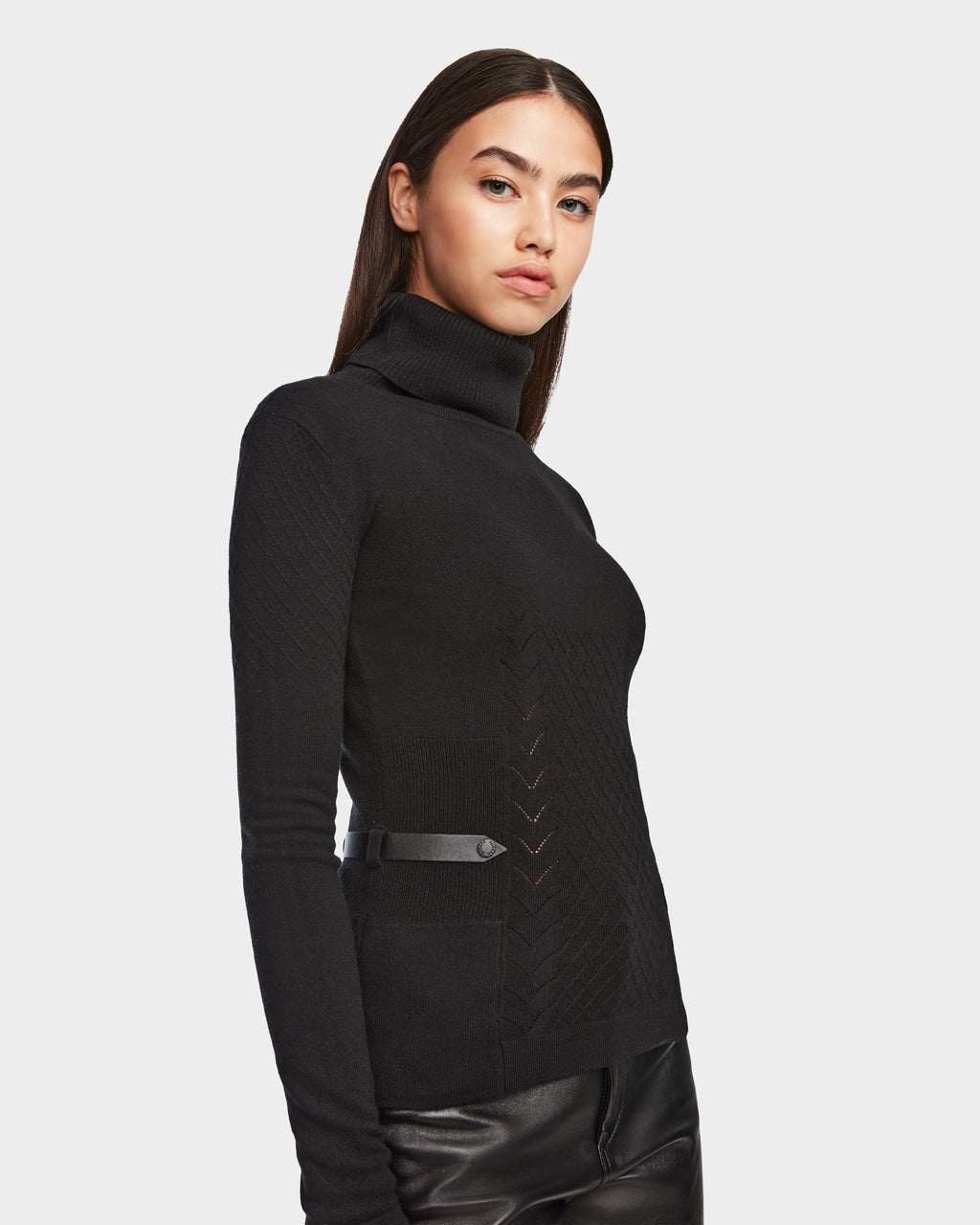 A woman with straight brown hair models the Rudsak LAURA - 8524596 Black womens knit turtleneck sweater with chevron detailing and vegan leather trim, paired with leather pants against a plain white background.