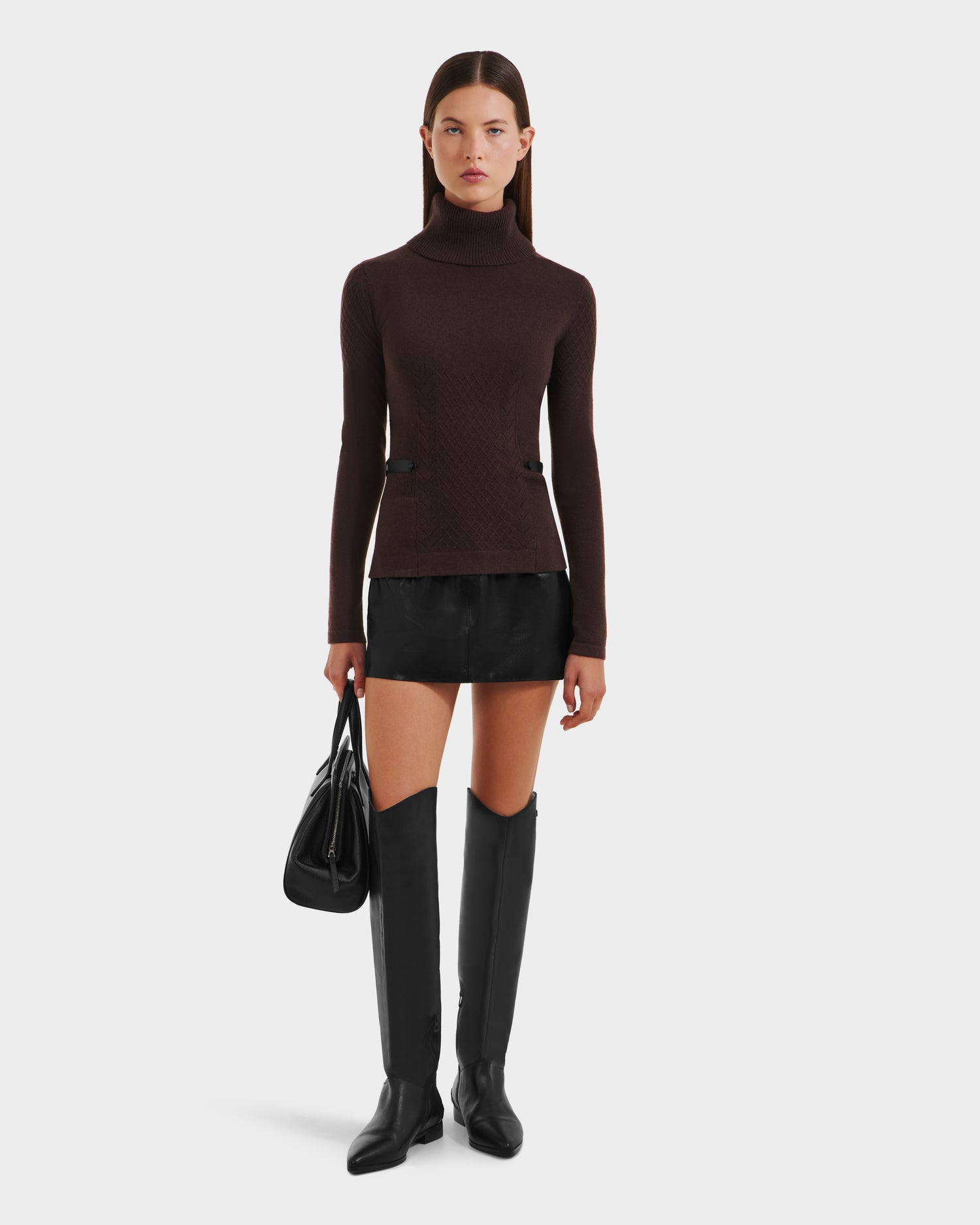 A woman stands facing forward wearing the Rudsak LAURA - 8524596 Mocha womens knit turtleneck sweater, paired with a black mini skirt, knee-high boots, and a black handbag with vegan leather trim. She has straight dark hair and a neutral expression.