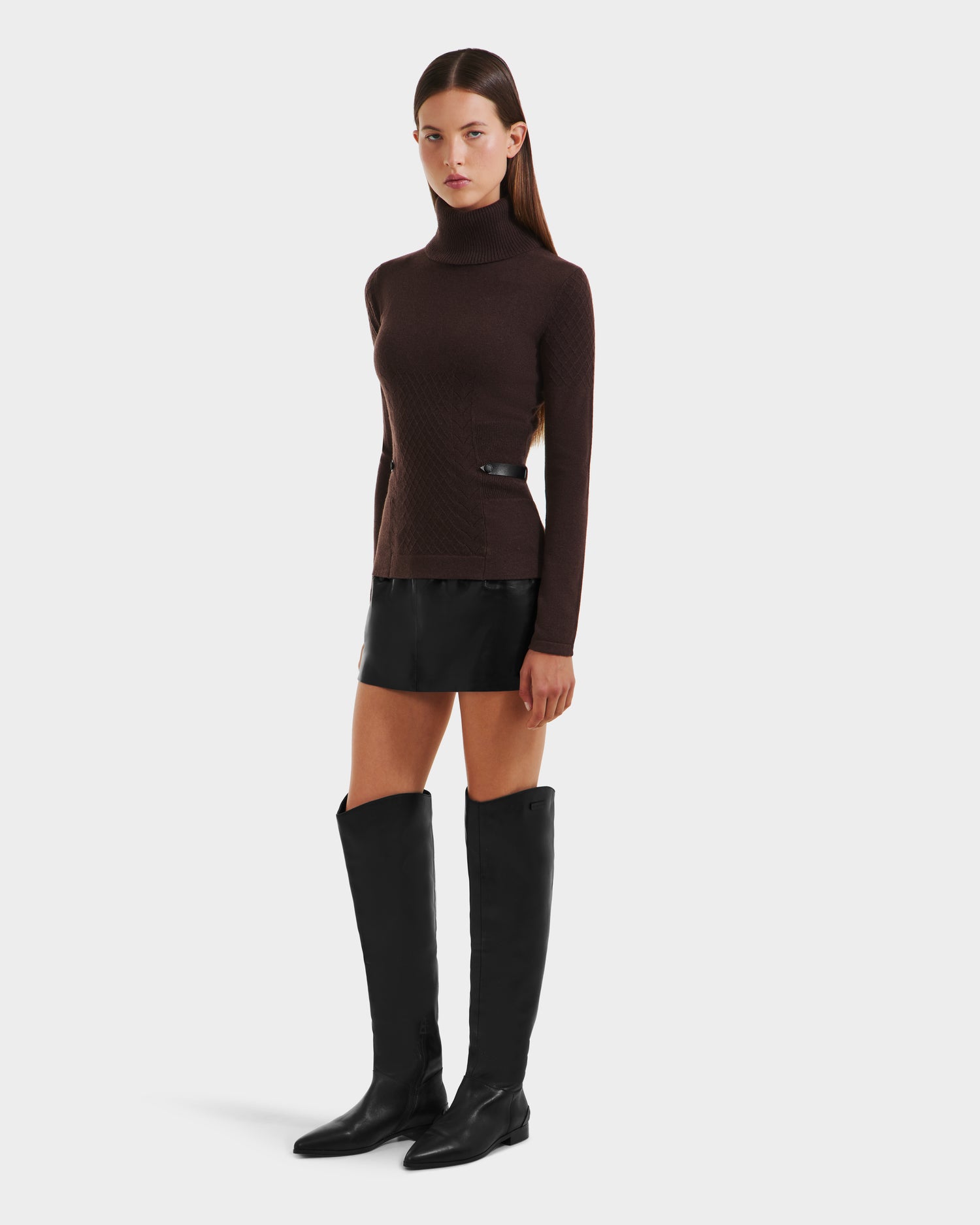 A woman with straight brown hair and a neutral expression stands against a plain background, wearing the Rudsak LAURA - 8524596 Mocha womens knit turtleneck sweater, a short black skirt with vegan leather trim, and tall black boots.