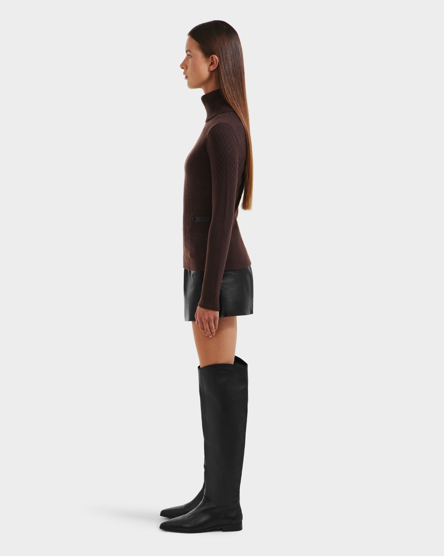 A woman with long brown hair stands in profile against a white background, wearing the Rudsak LAURA - 8524596 Mocha womens knit turtleneck sweater, a leather-trimmed black mini skirt, and knee-high black boots.